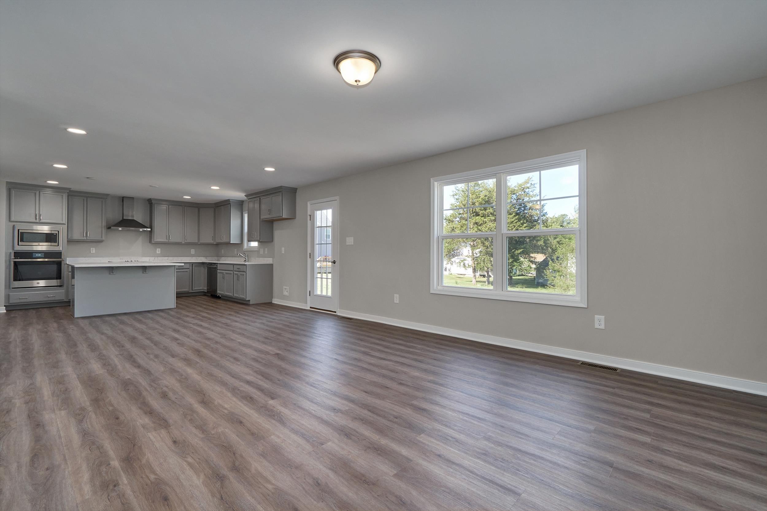An empty, spacious open-concept kitchen and living area with gray cabinets, hardwood flooring, and large windows providing natural light.
