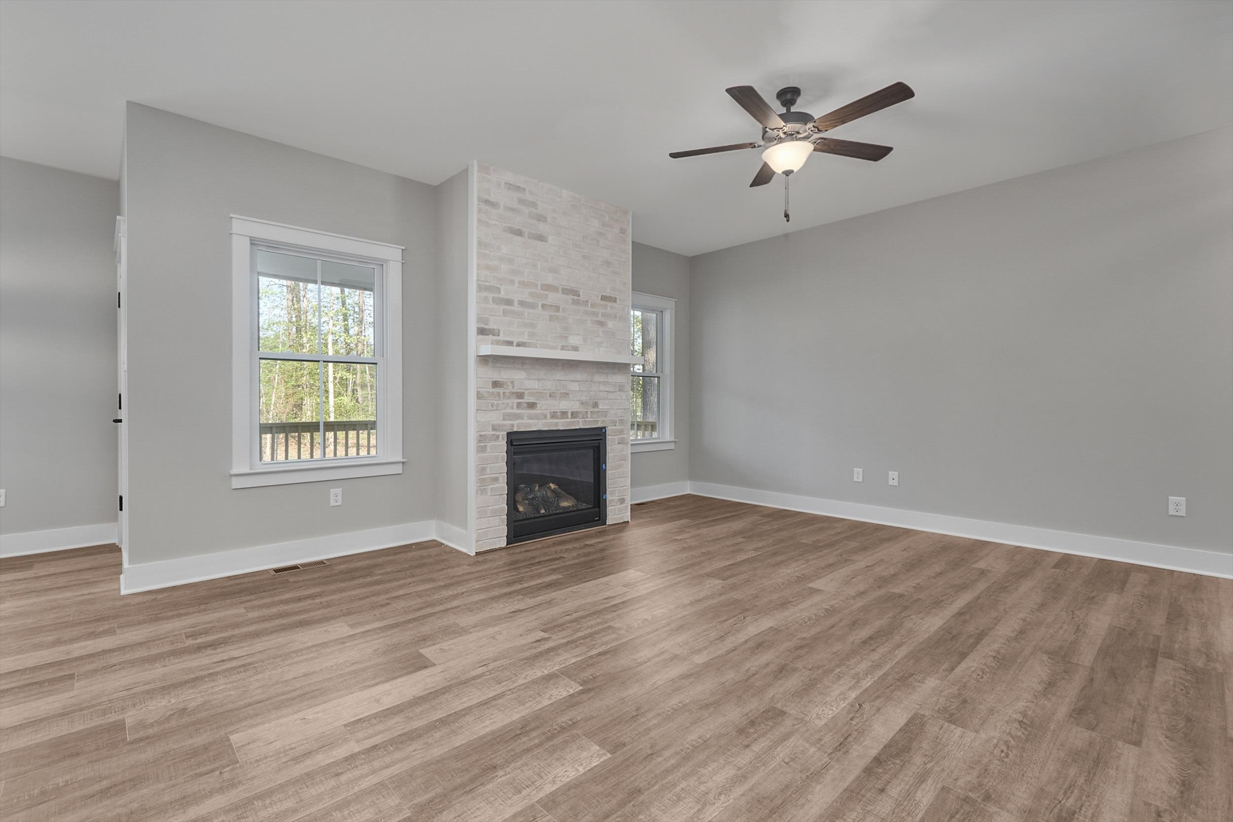 Spacious living room with light gray walls, a wooden floor, a ceiling fan, and a fireplace framed by two windows.