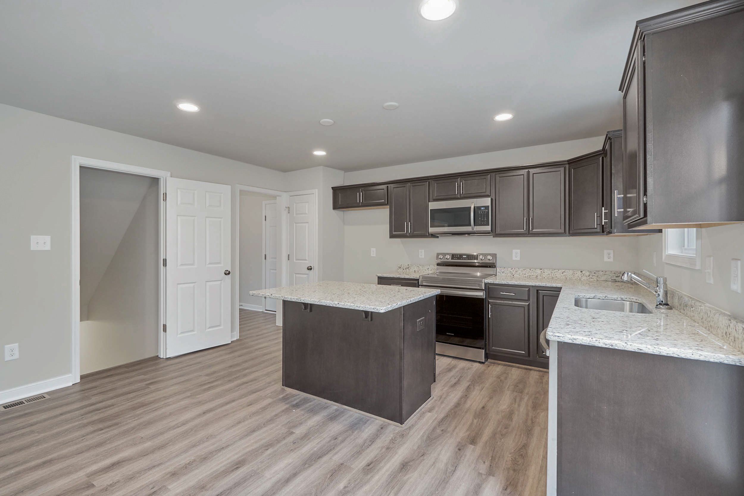Modern kitchen with dark wood cabinets, stainless steel appliances, a central island, light granite countertops, and wood flooring. White doors are visible in the background.