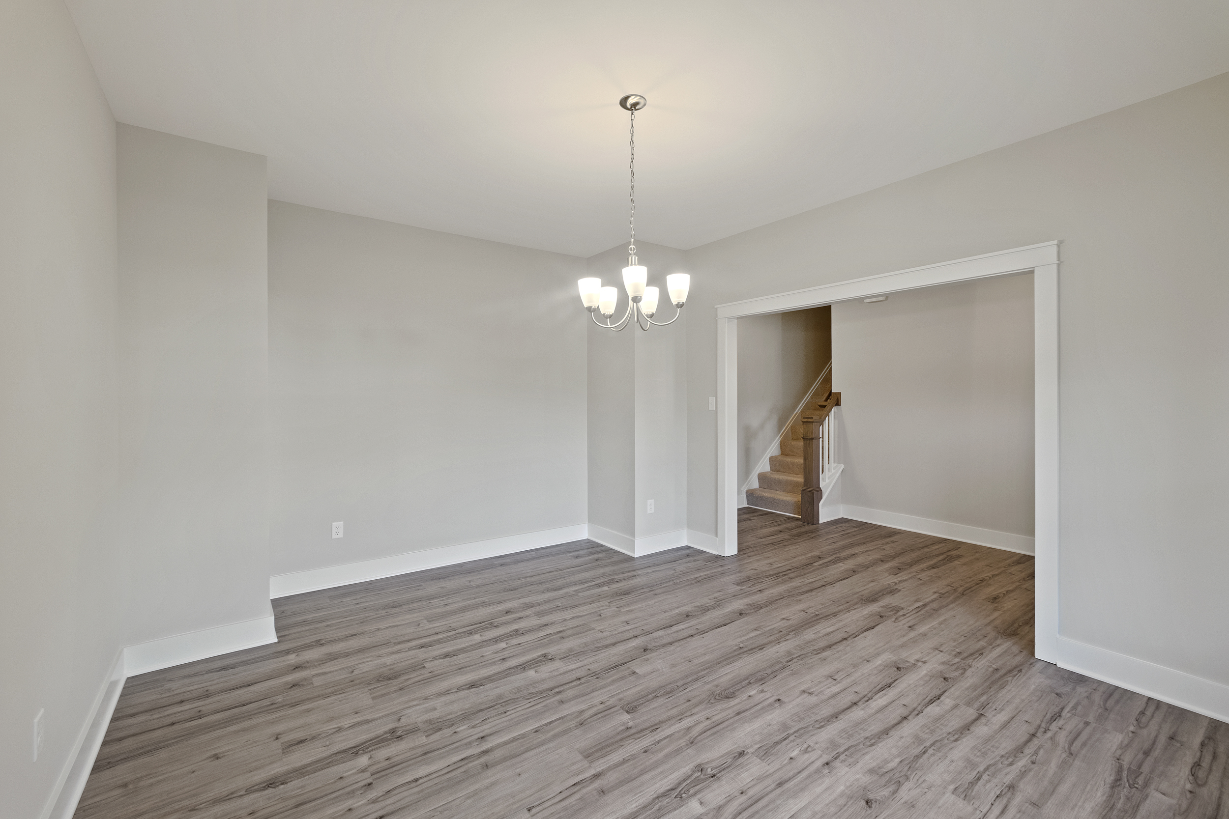 A minimalist room with light gray walls, wood flooring, a modern chandelier, and an open entryway leading to a staircase.
