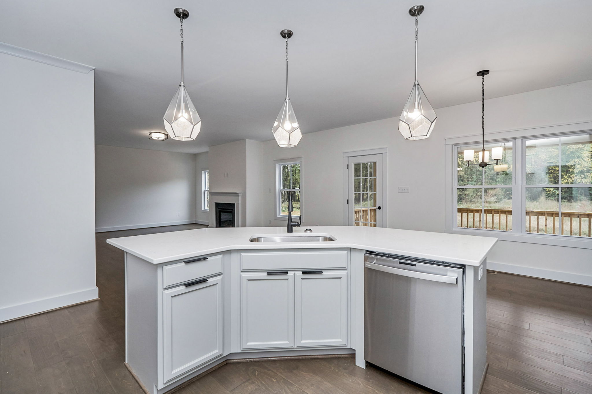 A modern kitchen with a white island featuring a sink and built-in dishwasher. Three geometric pendant lights hang above. In the background is the living area and a door leading outside.