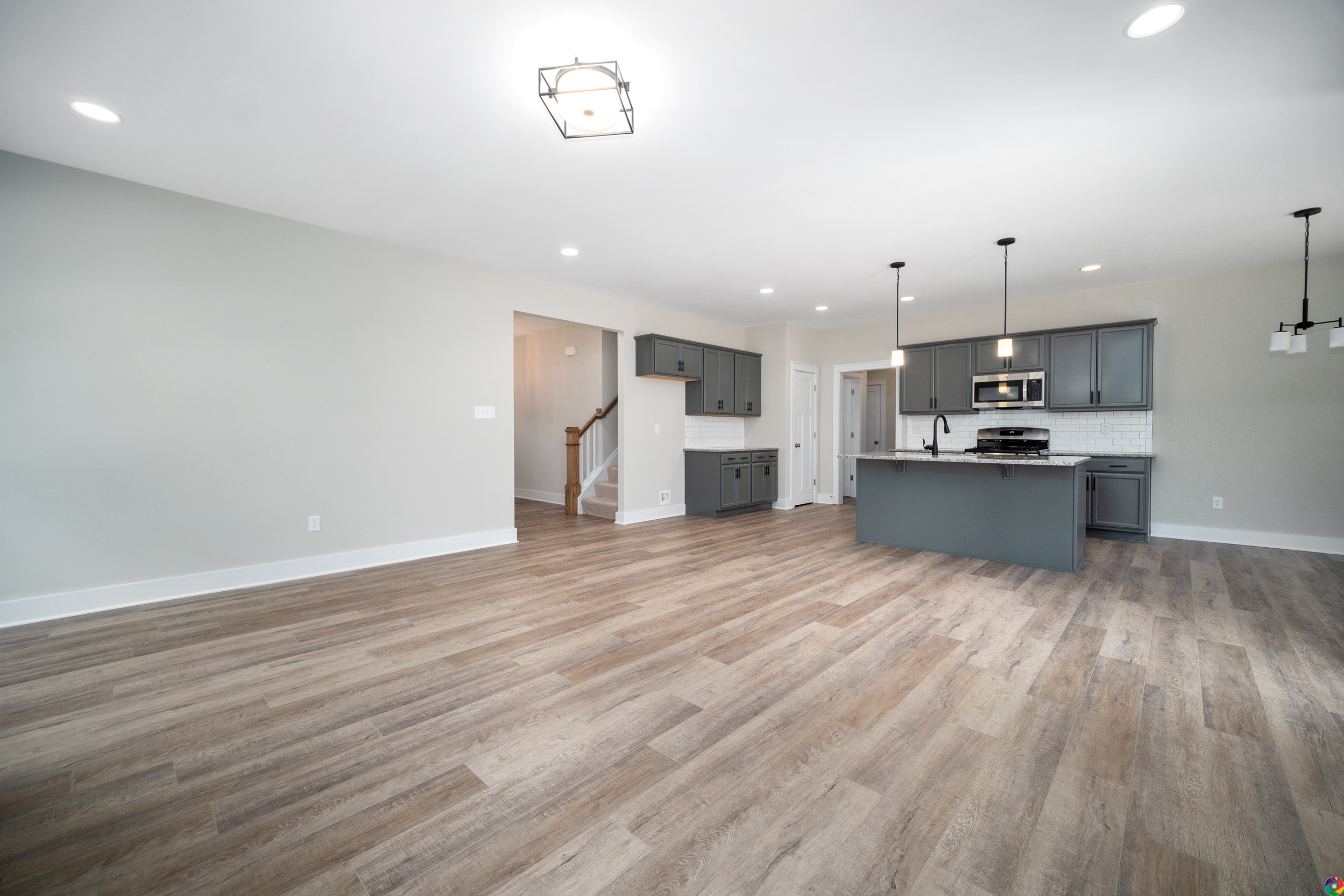 Modern kitchen and living area with hardwood flooring, grey cabinets, stainless steel appliances, and recessed lighting. Open floor plan includes a staircase in the background.