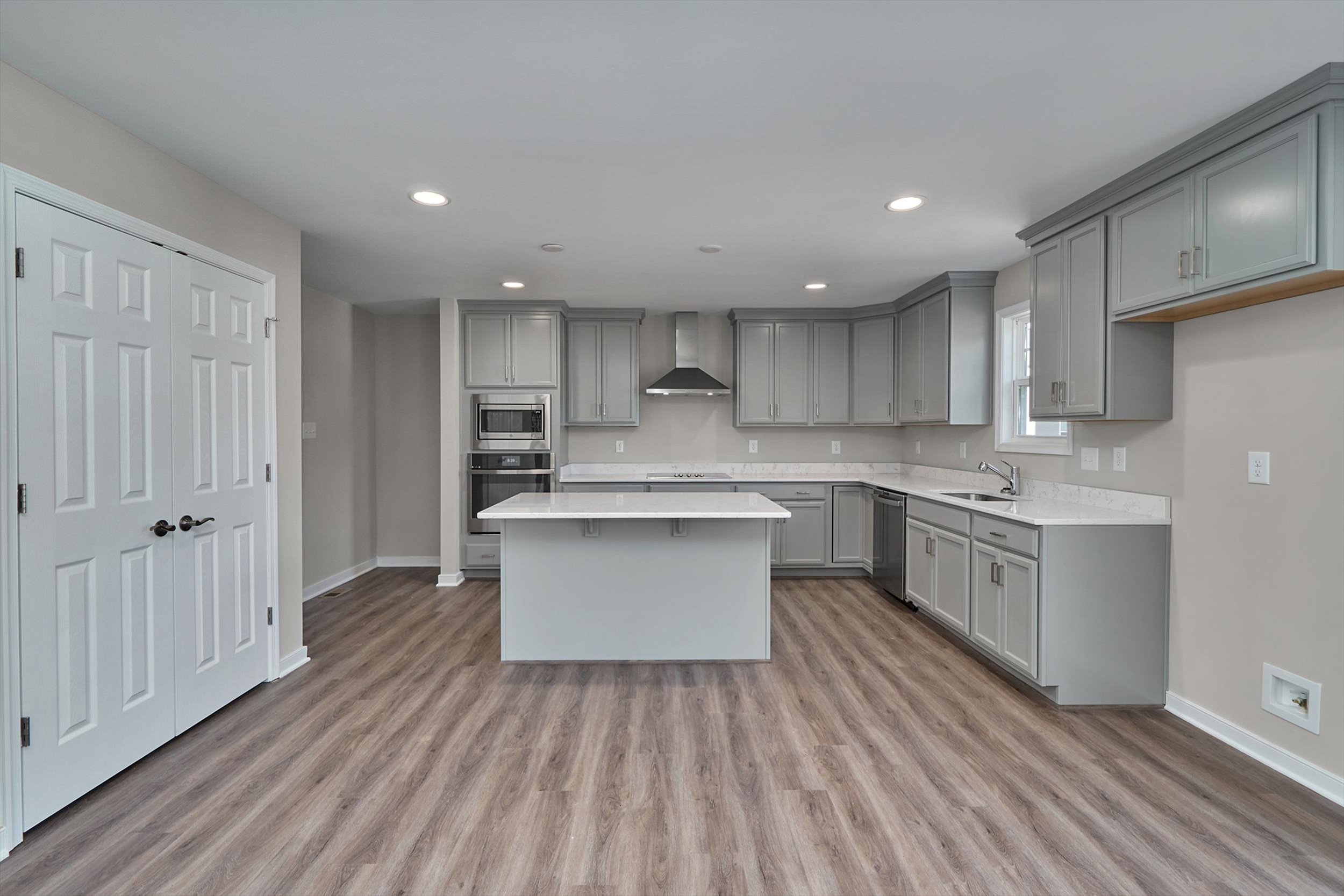 A modern kitchen with light gray cabinets, a central island, stainless steel appliances, wooden flooring, and recessed lighting.