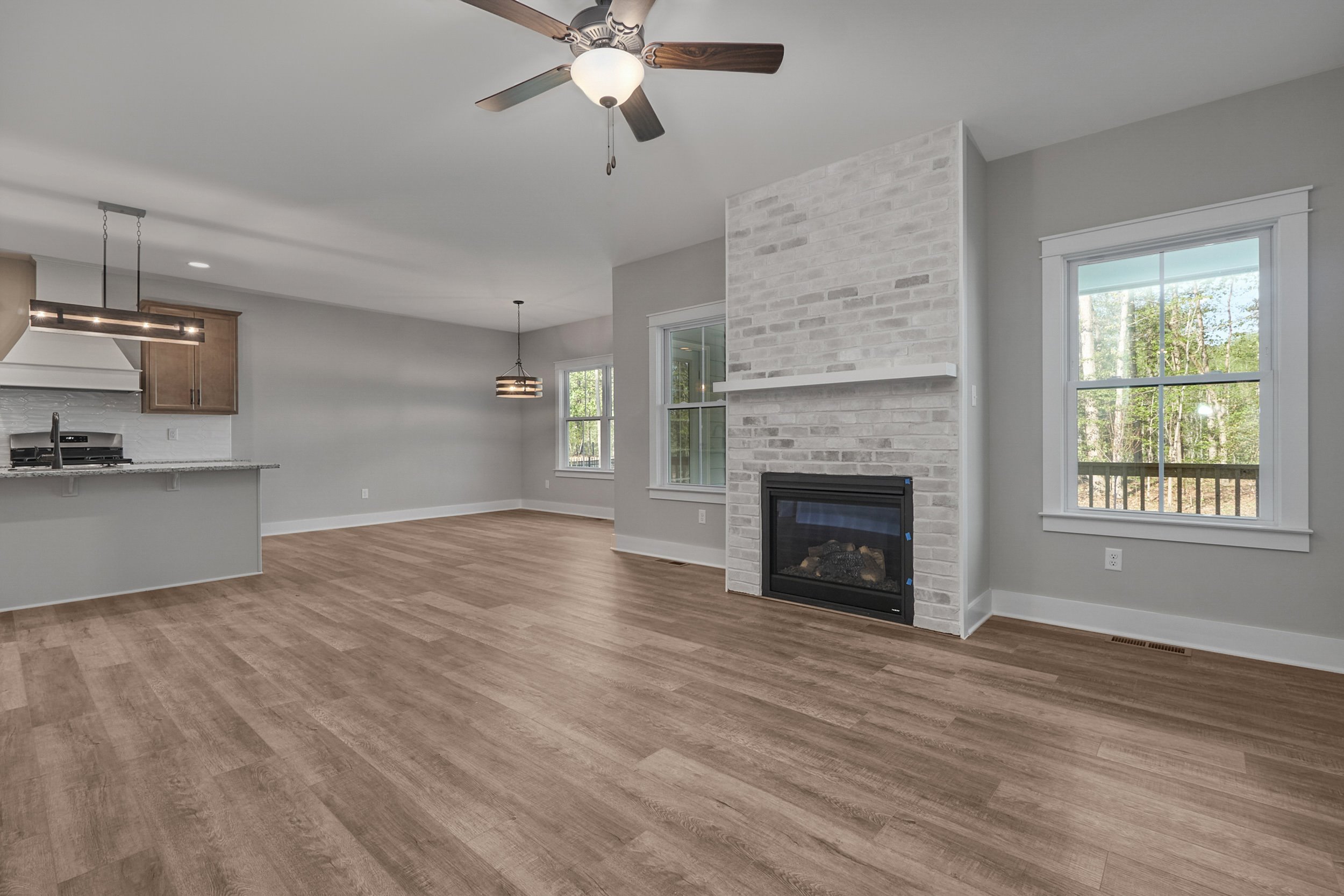 A spacious, empty living room with light wood flooring, a white brick fireplace, ceiling fan, and large windows. The room opens to a kitchen with an island and modern light fixtures.