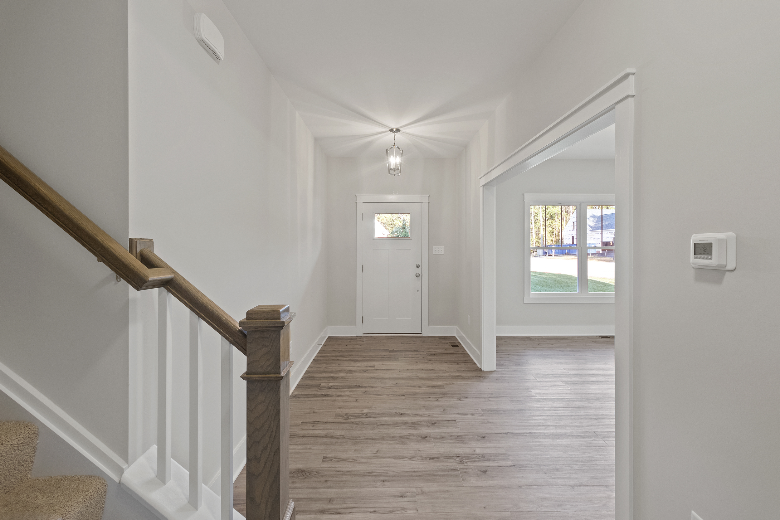 A well-lit foyer with a light fixture, a white front door, wooden floors, a staircase with white railings, and an adjacent room with large windows visible.