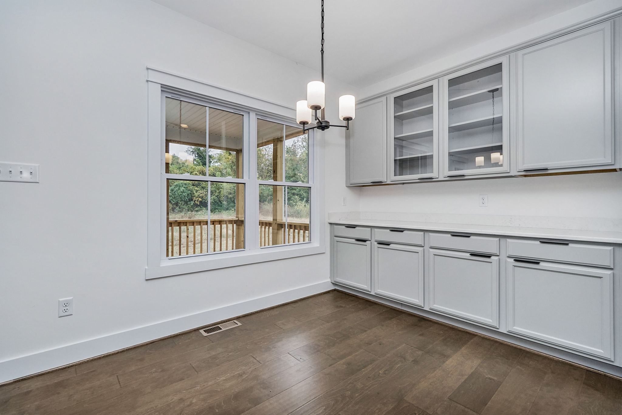 A kitchen area with wooden floors, gray cabinets, a chandelier light fixture, a large window overlooking a deck, and a white countertop beneath glass-fronted cupboards.
