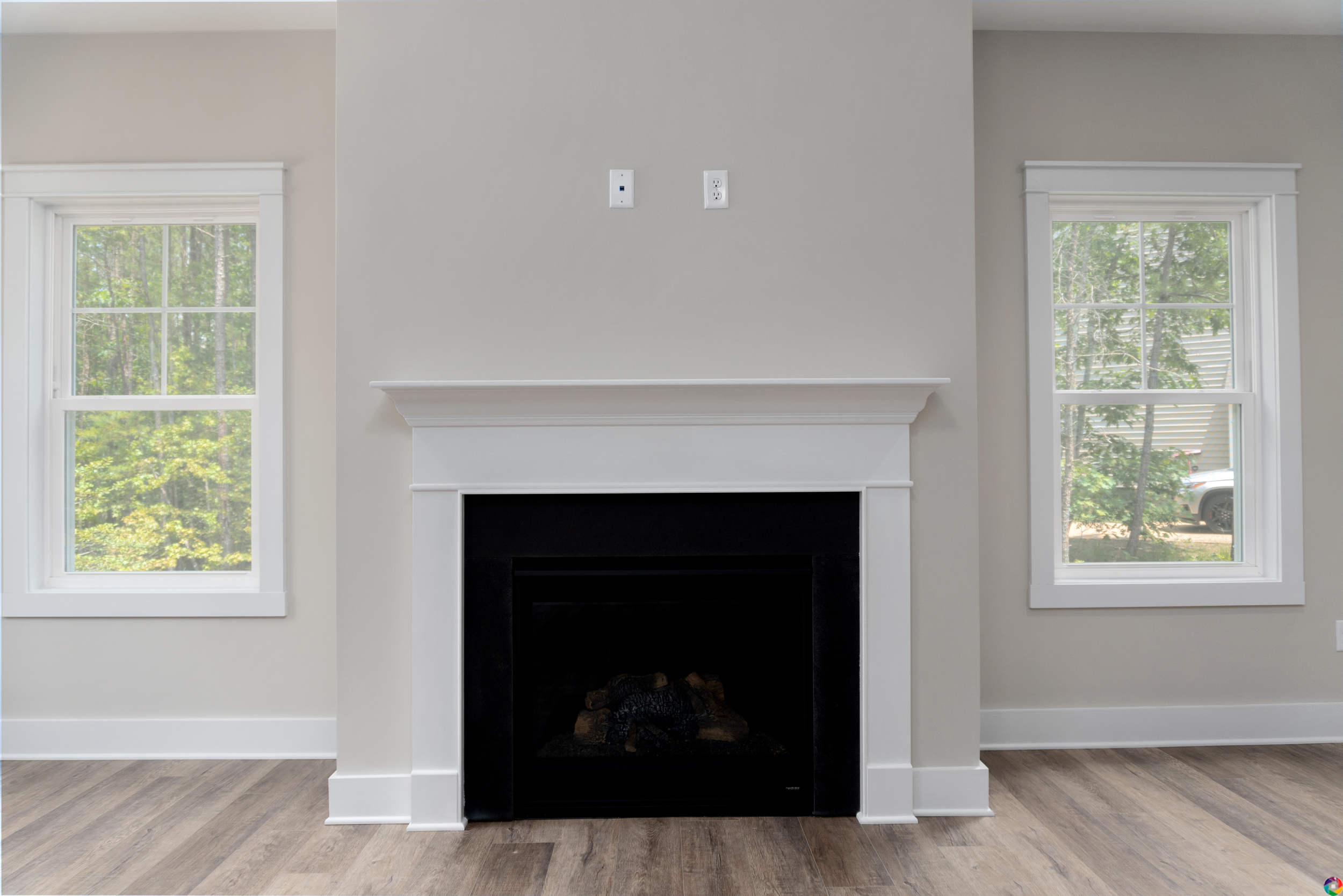 A modern fireplace with a white mantel is centered between two windows in a room with light gray walls and wood flooring.
