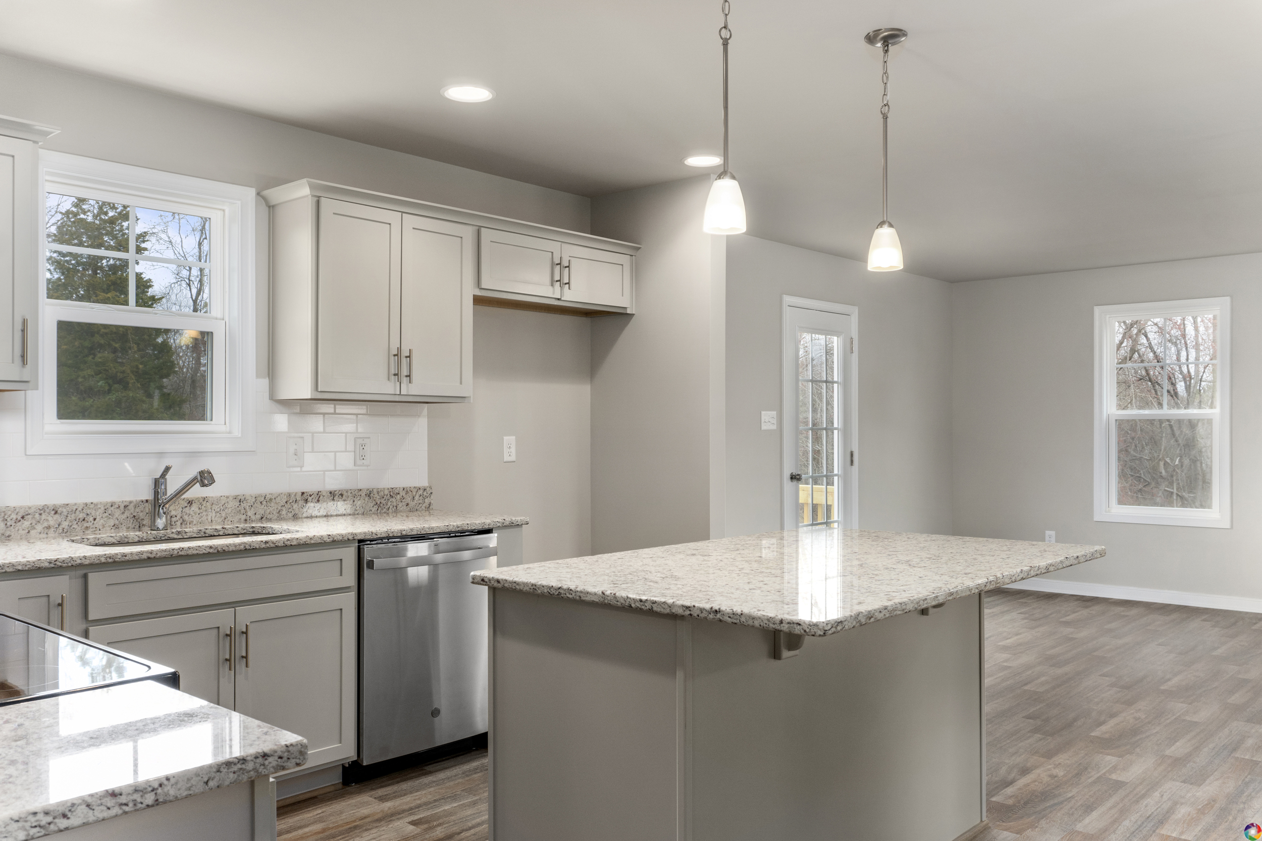 A modern kitchen with gray cabinets, granite countertops, stainless steel appliances, two pendant lights, and a central island. A door and windows allow natural light into the space.