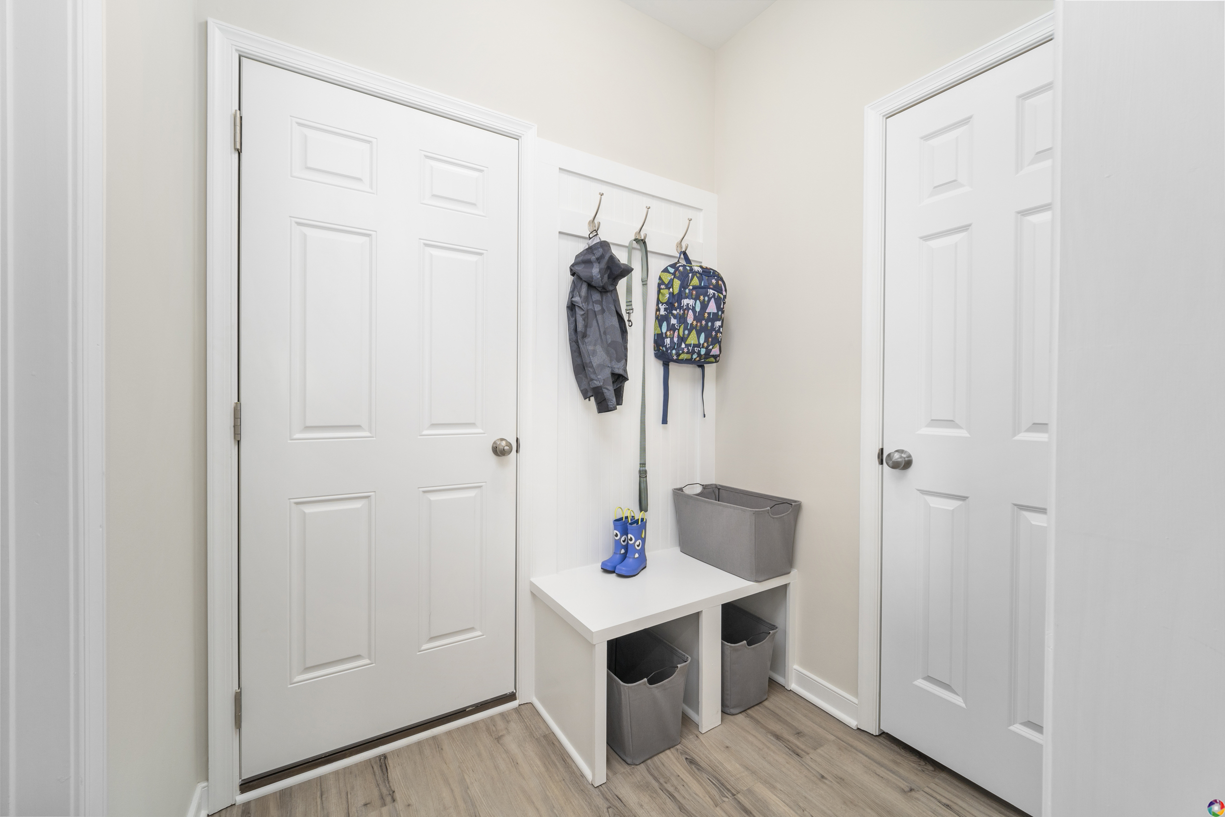 A small, tidy entryway with two white doors, a bench with storage cubes underneath, and hooks holding a jacket and a backpack.