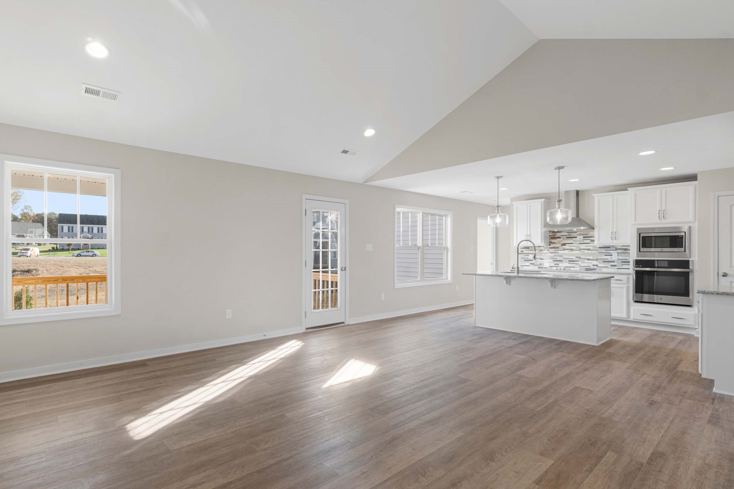 Modern kitchen with white cabinets and a central island opening to a spacious, sunlit living room with large windows and wooden flooring.
