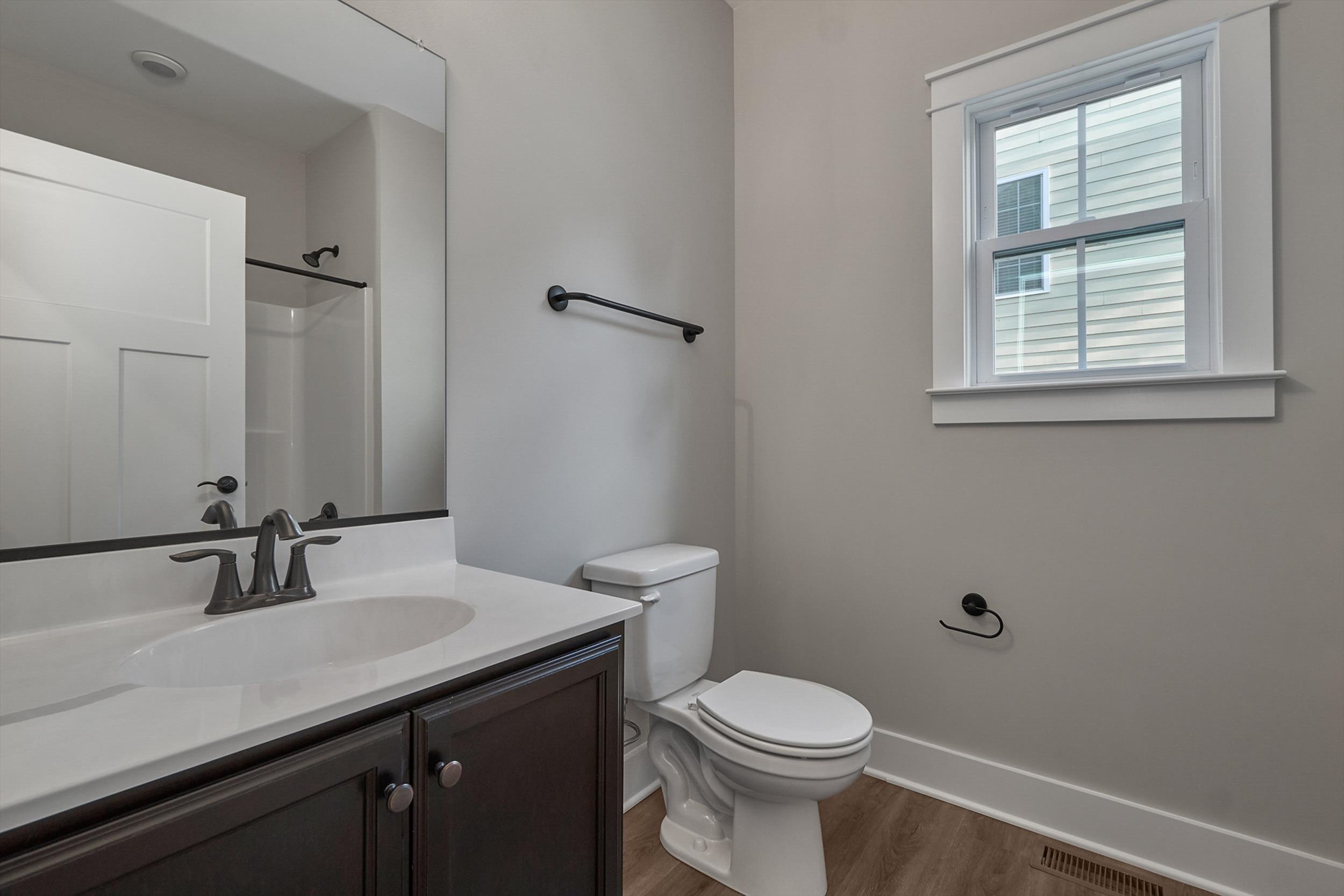 A bathroom with a sink, faucet, mirror, toilet, and a window. Walls are painted light gray. The floor is wooden, and there's a white door leading to a shower area.