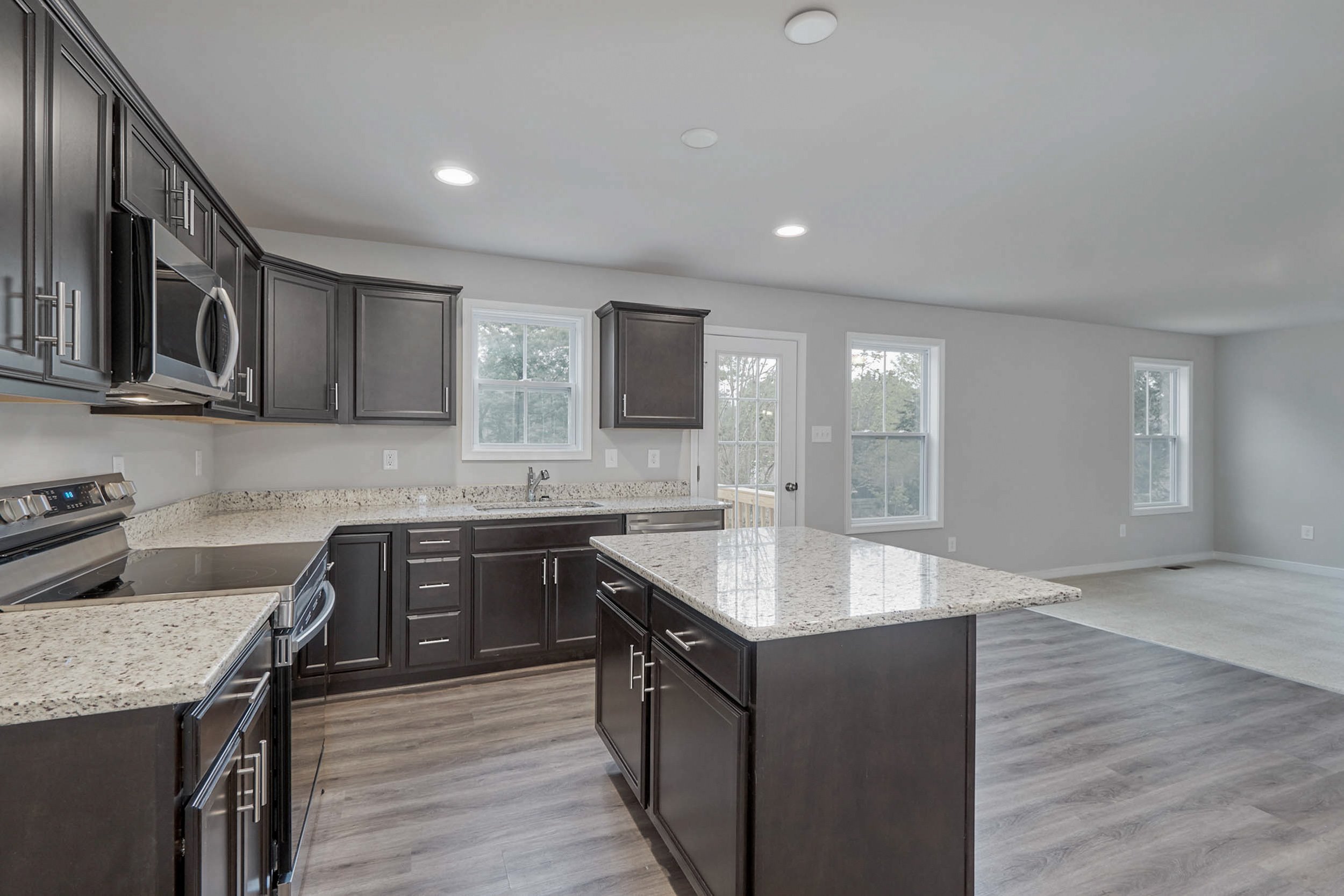 A modern kitchen with dark cabinetry, granite countertops, an island in the center, stainless steel appliances, and wooden flooring. Bright natural light enters through multiple windows.