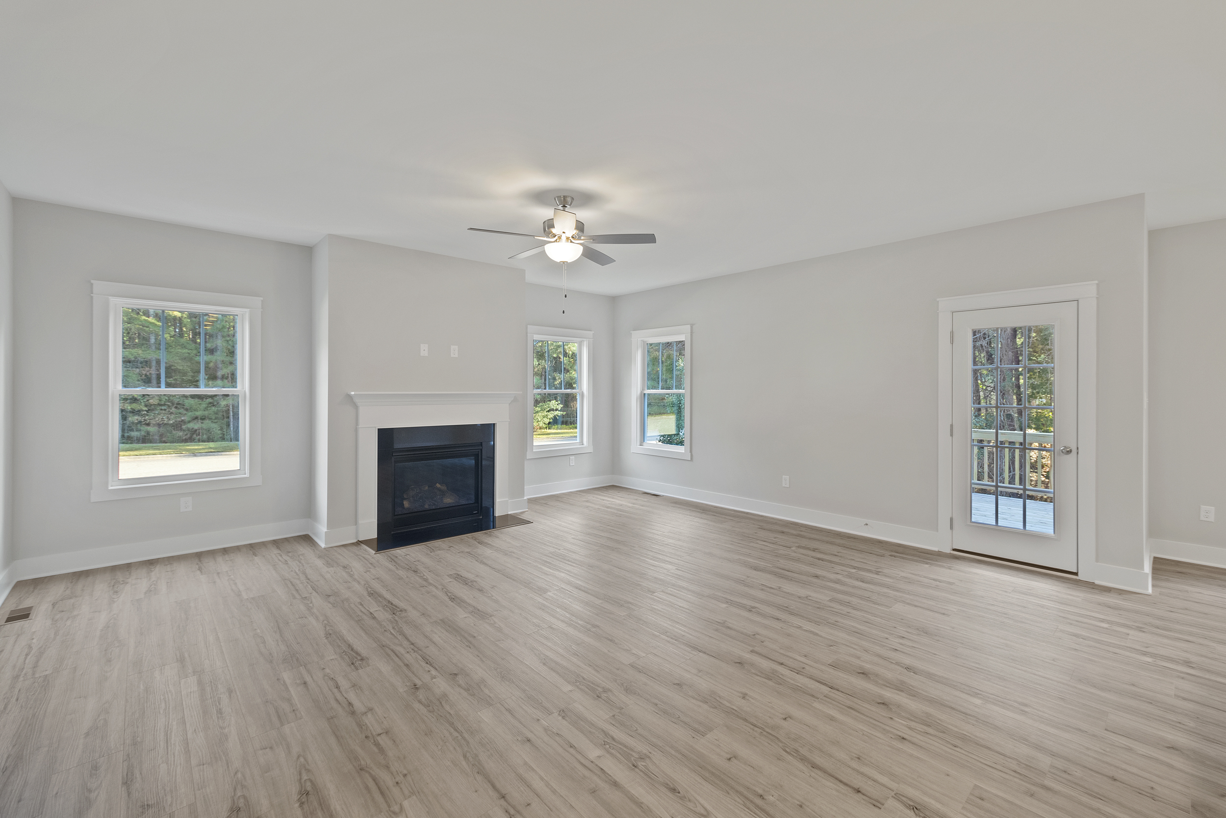 A spacious, empty living room with light wood flooring, white walls, a ceiling fan, a fireplace, three windows, and a glass door leading to an exterior balcony.