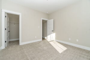 Empty bedroom with beige walls, carpet flooring, and sunlight streaming through a window. two open doors reveal closet space.
