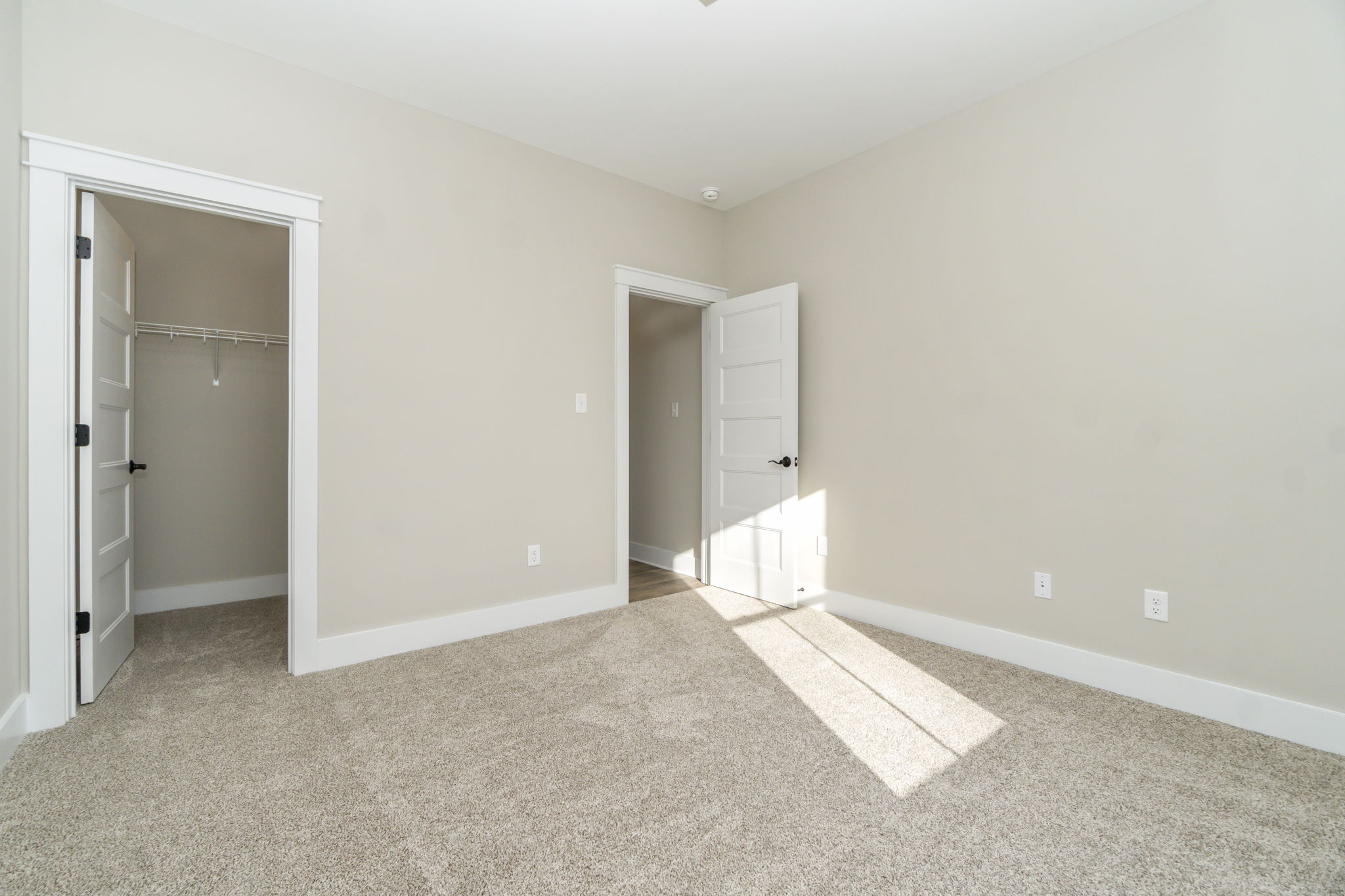 Empty bedroom with beige walls, carpet flooring, and sunlight streaming through a window. two open doors reveal closet space.