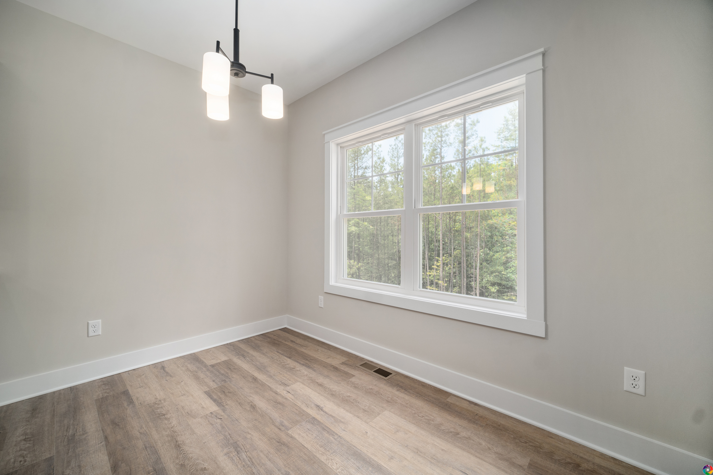 A minimalist room with light wood flooring, a large window overlooking trees, and a modern ceiling light fixture with three shades. Walls are painted light gray. Room is empty.