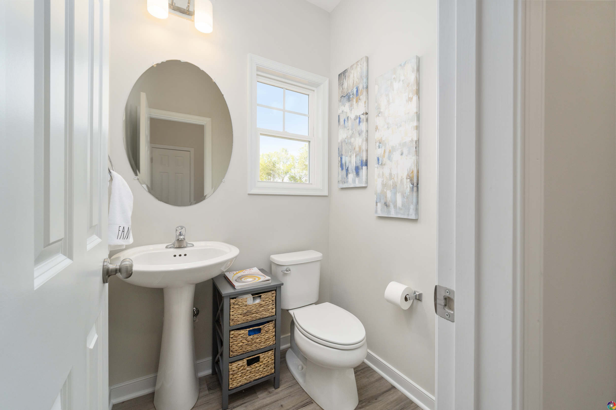 A small bathroom with a round mirror above a pedestal sink, a toilet, a window, a towel ring, and two abstract paintings on the wall. There is a storage unit with wicker baskets beside the sink.