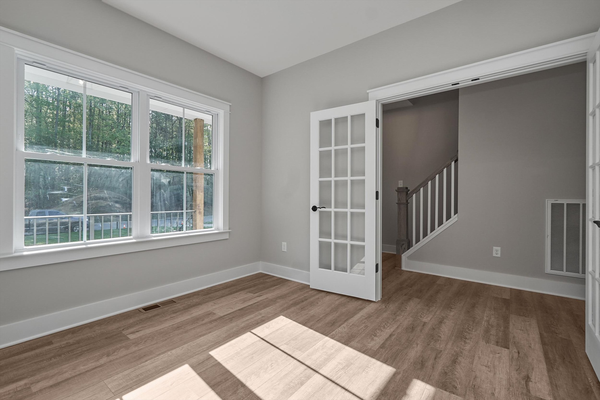 A room with large windows, French doors, light-colored walls, and hardwood flooring, adjacent to a hallway with a staircase. Plenty of natural light enters through the windows.