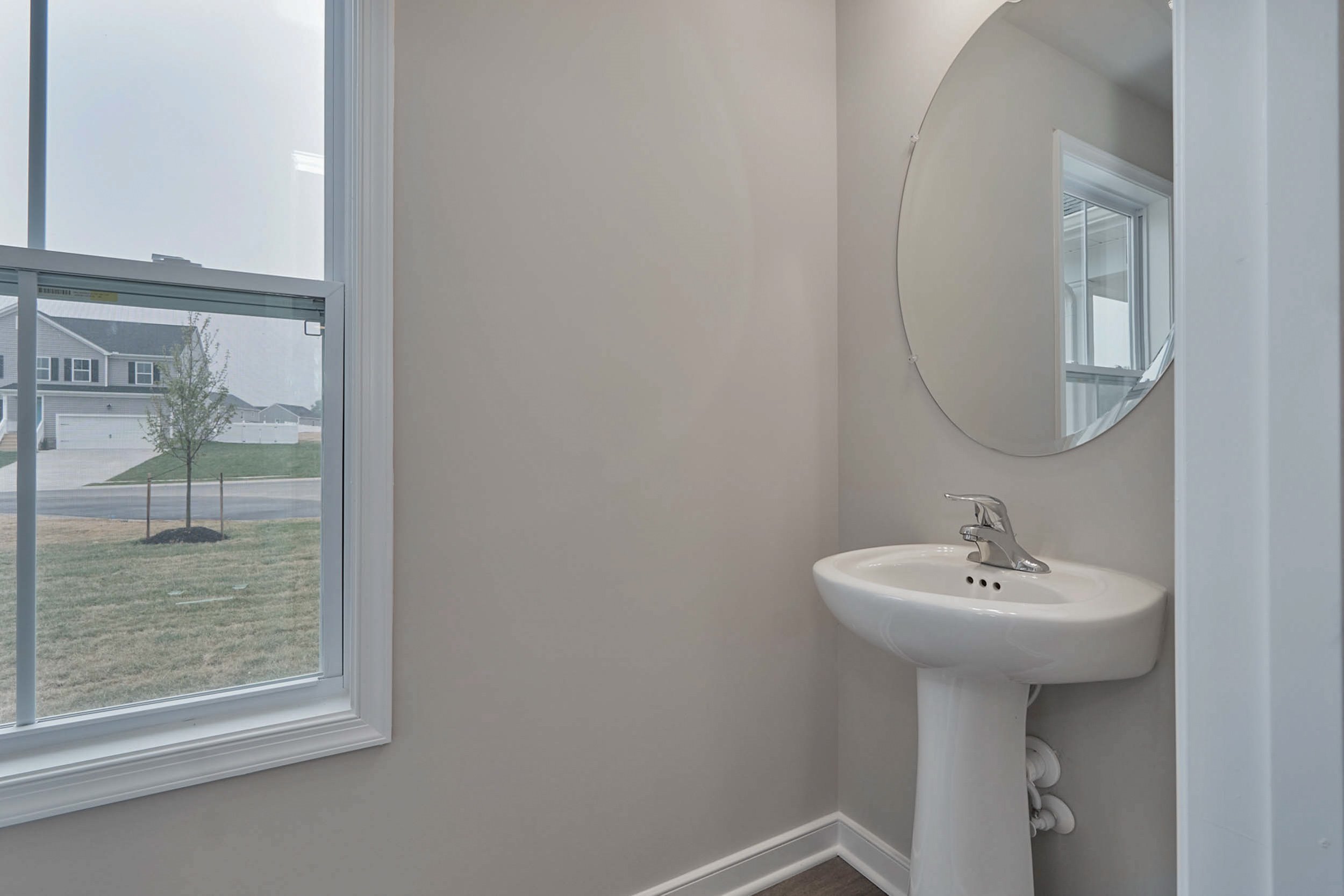A small bathroom with a pedestal sink, round mirror, and a window showing a view of the outside. The walls are painted light gray.