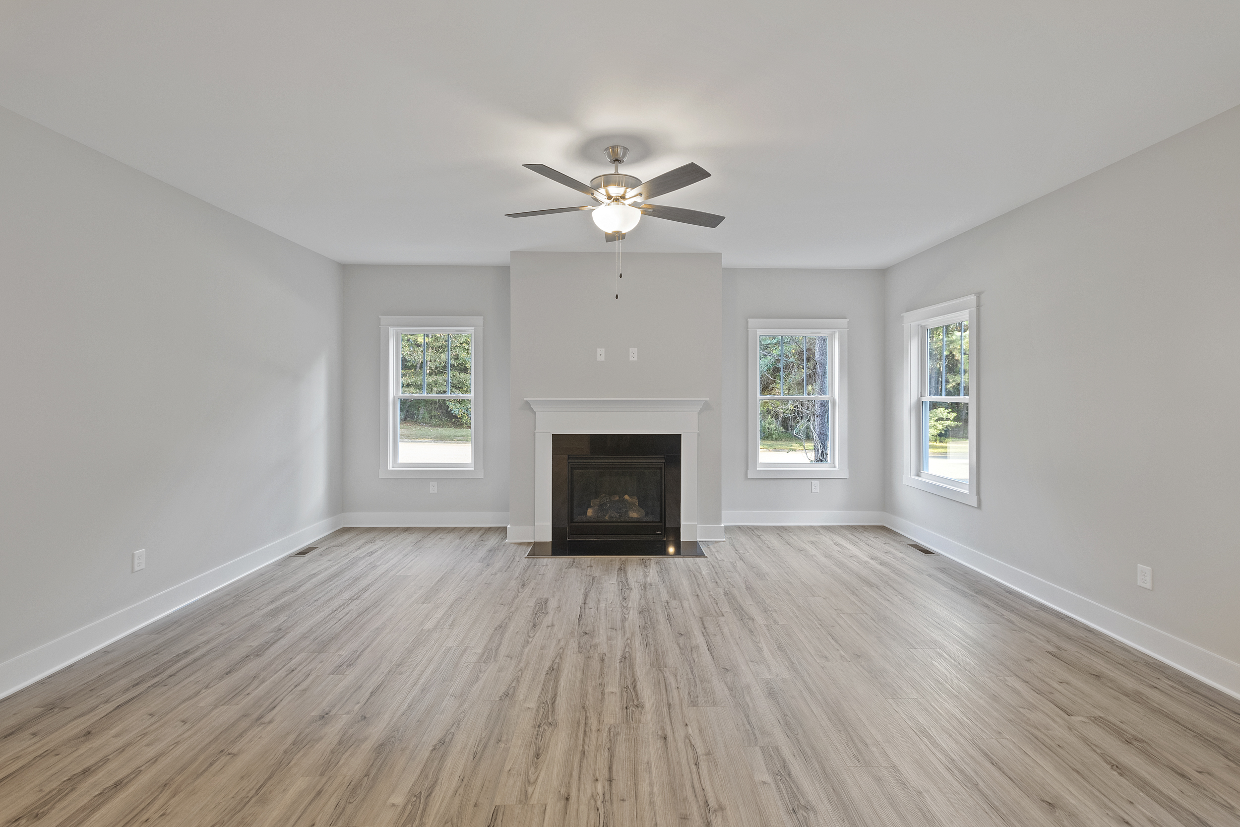 A spacious, empty living room with light wood flooring, a ceiling fan, a fireplace with a white mantle, and three large windows offering natural light.