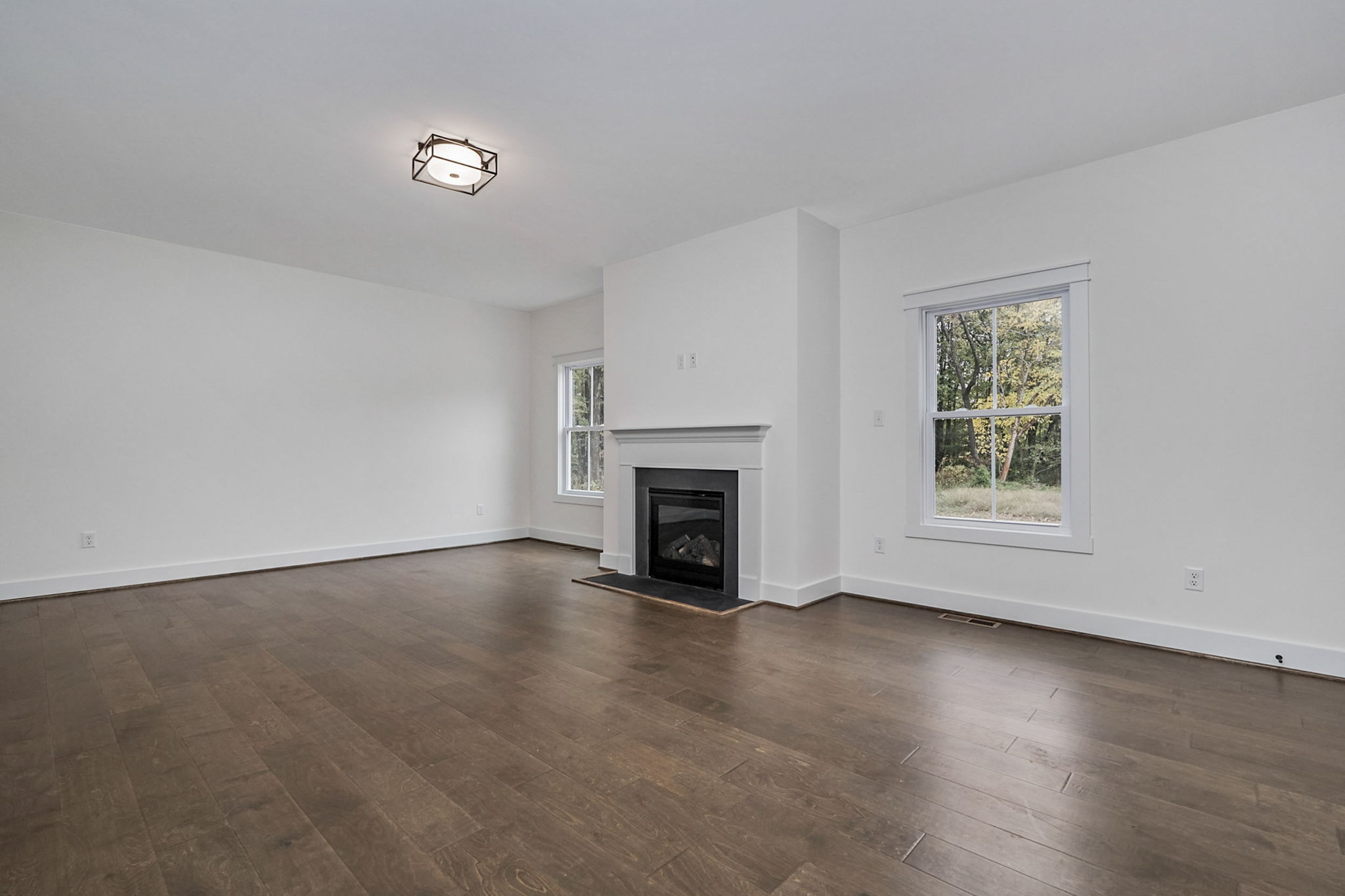 An empty room with hardwood flooring, white walls, a ceiling light fixture, a fireplace against one wall, and two windows providing natural light.