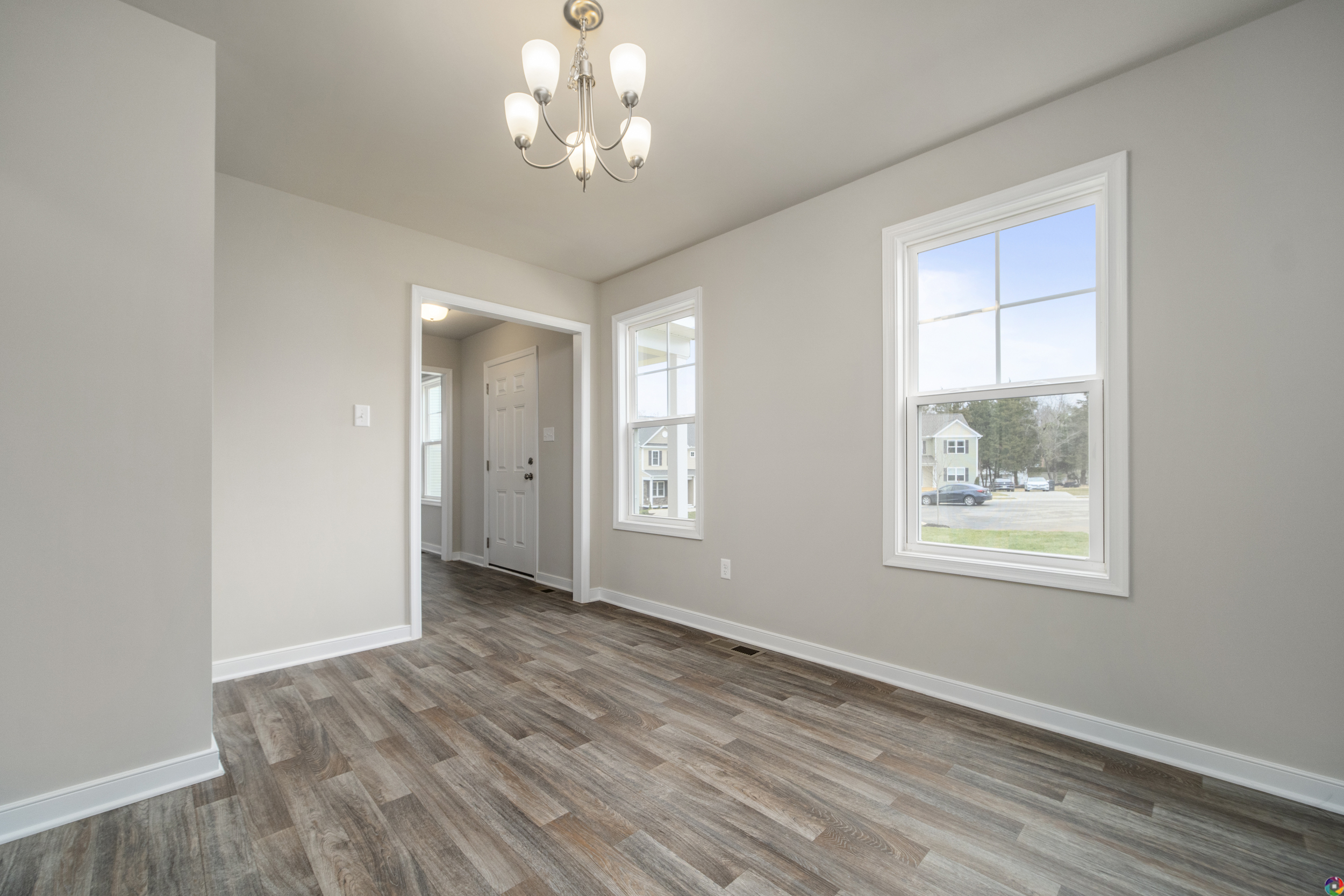 An empty room with gray walls, two windows, laminate wood flooring, and a chandelier. A doorway leads to an adjacent hallway and an exterior door.