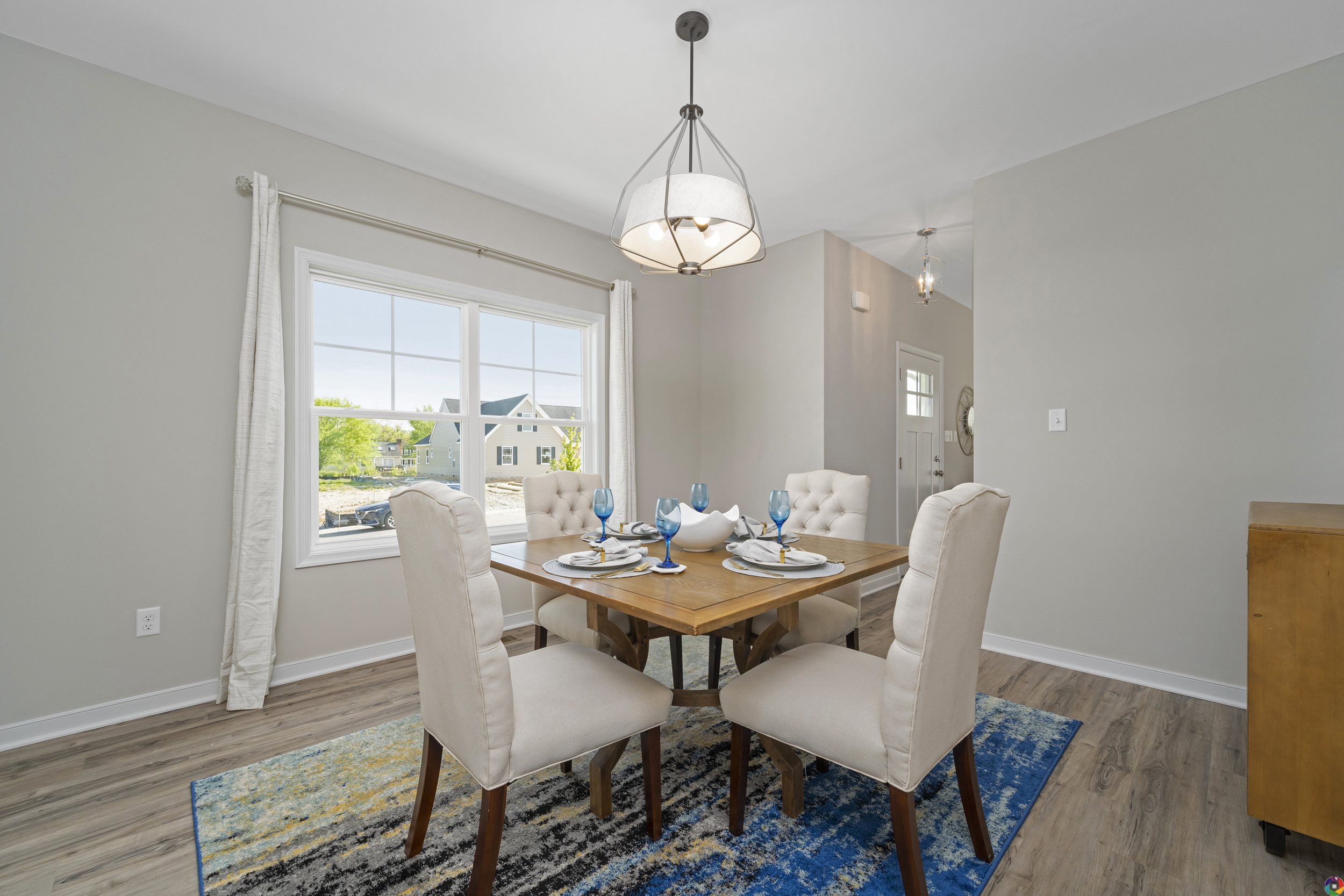 A bright, modern dining room featuring a round table set for four, beige chairs, a colorful area rug, and an elegant pendant light, with a large window showcasing a suburban view.