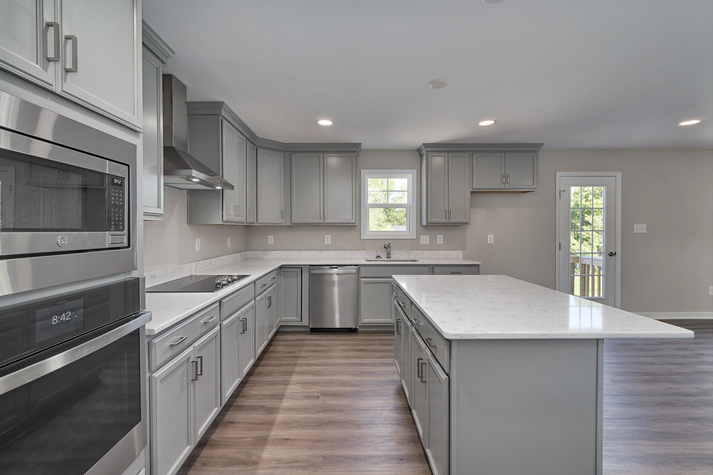 Modern kitchen with grey cabinets, stainless steel appliances, a large island with a white marble countertop, and hardwood floors. A window and glass door bring natural light into the room.