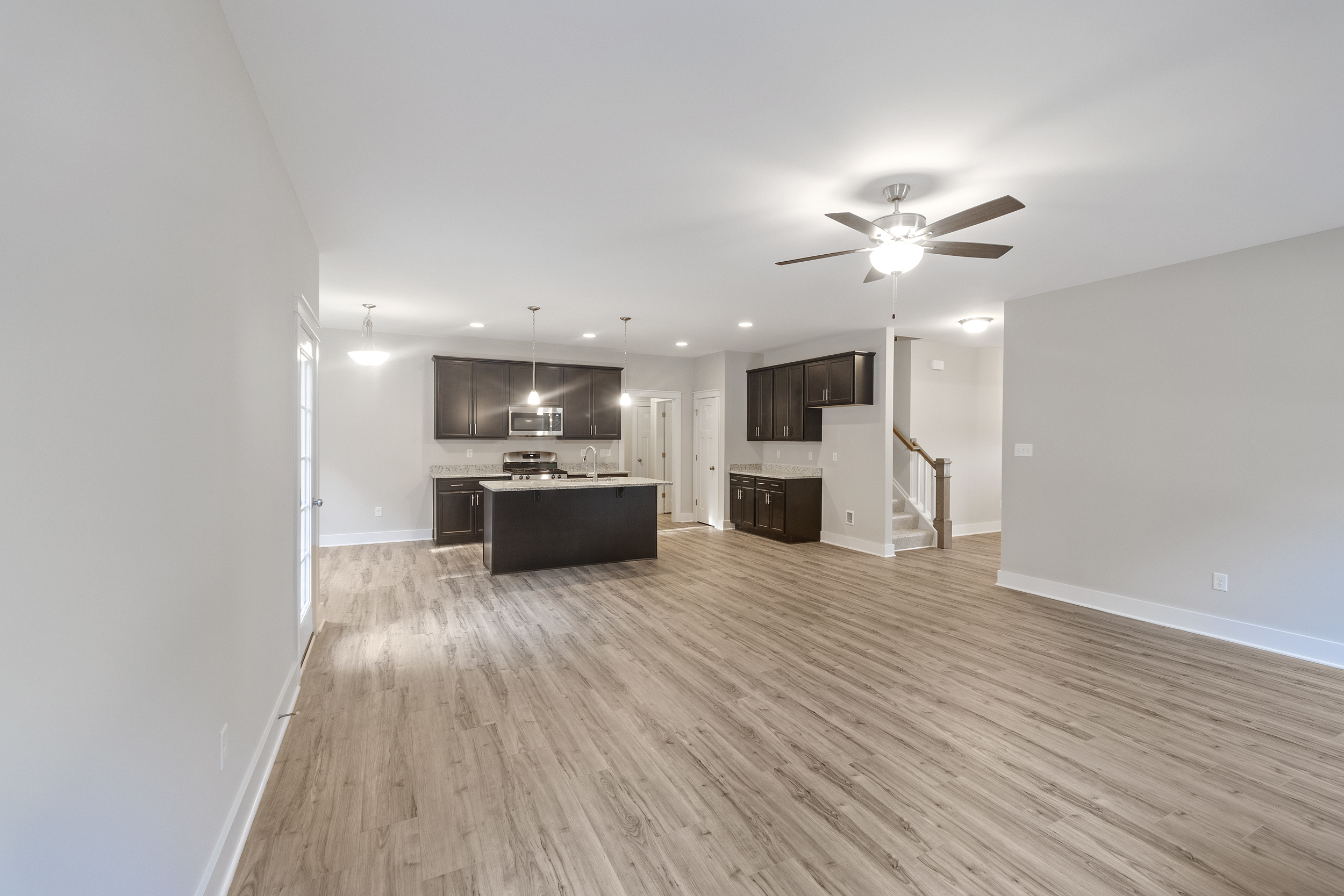 An empty, modern open-concept living room and kitchen with wooden floors, a ceiling fan, and recessed lighting. The kitchen has dark cabinets, an island, and stainless steel appliances.
