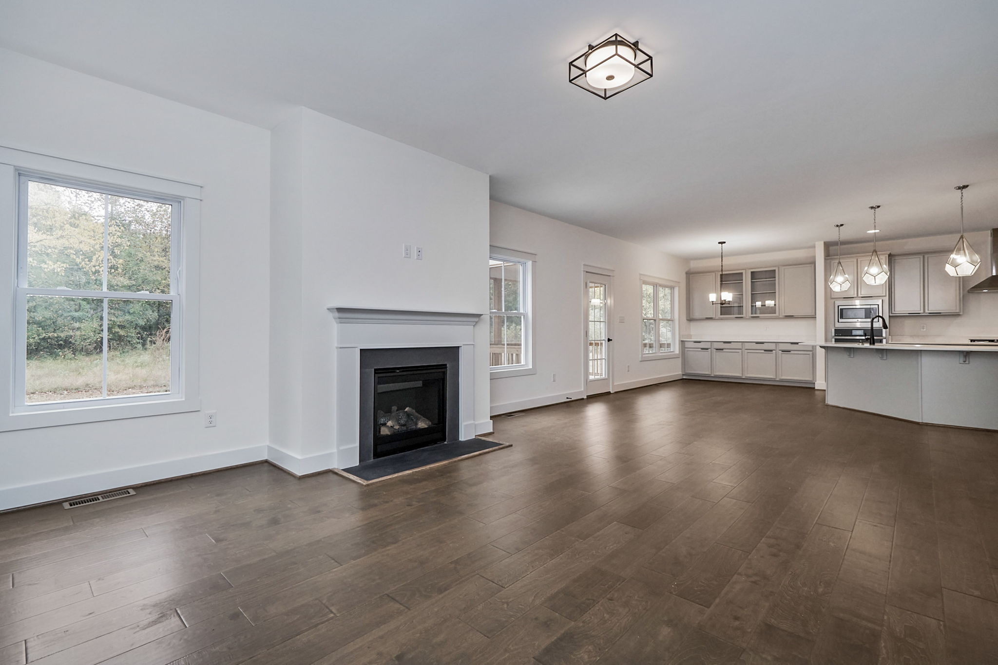Spacious open-concept living area with hardwood floors, a fireplace, modern lighting fixtures, and a large kitchen featuring white cabinets, an island, and multiple pendant lights.