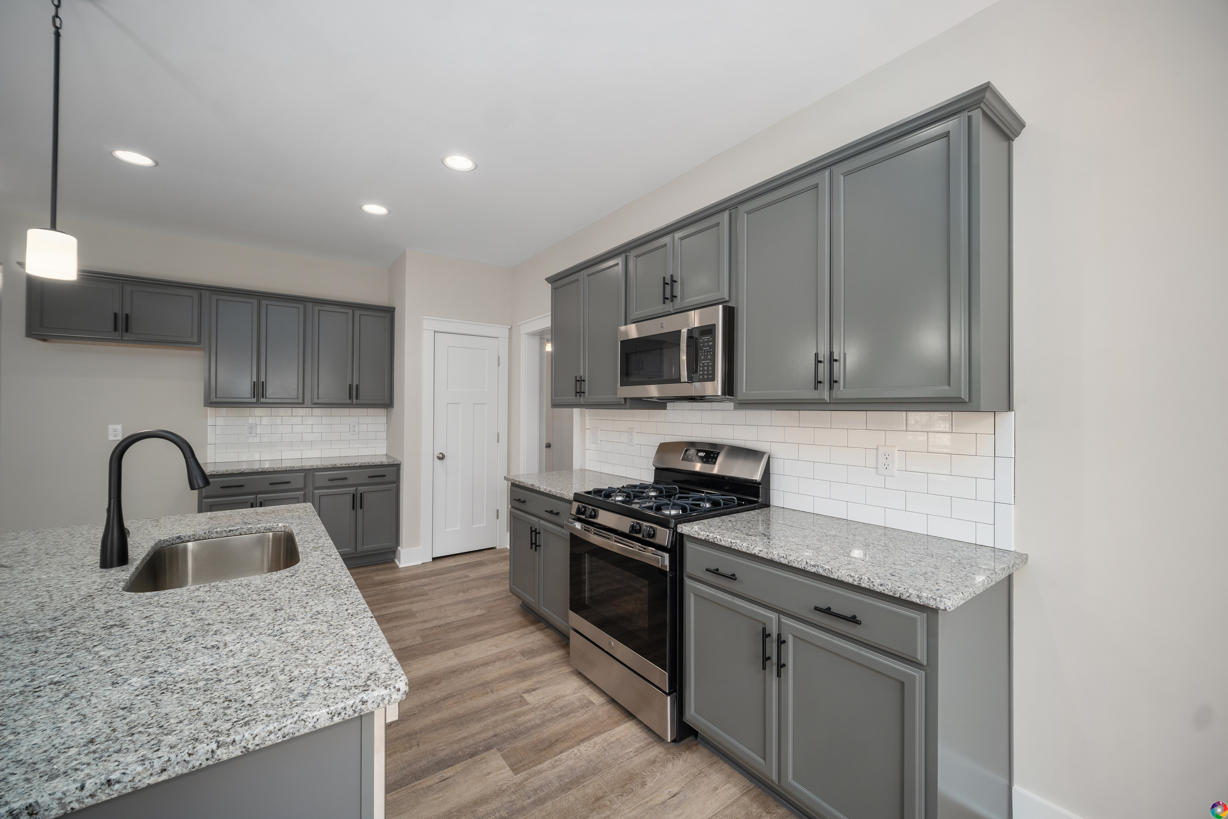 A modern kitchen featuring gray cabinets, granite countertops, a large island with a sink, stainless steel appliances, and a white subway tile backsplash.