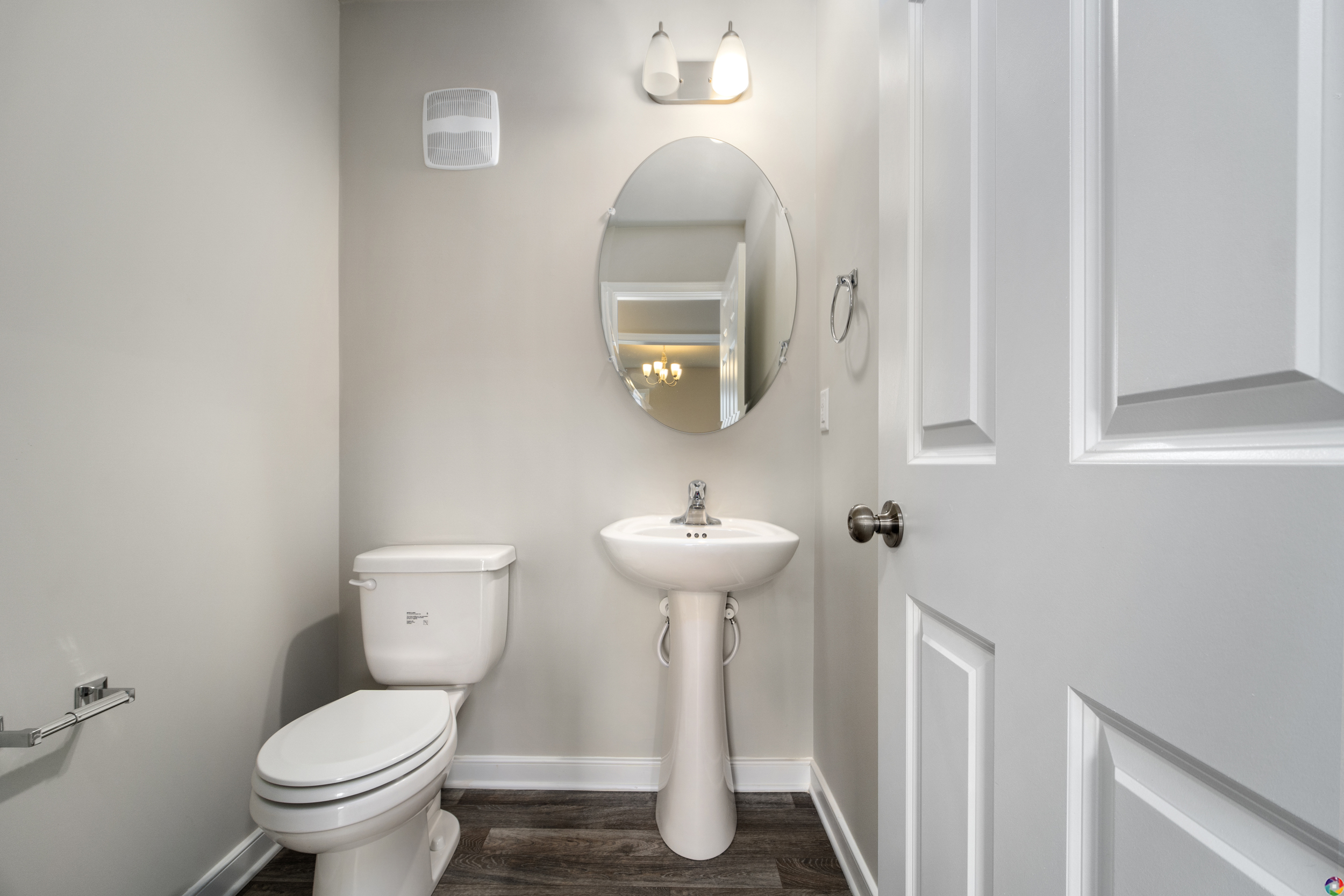 A small bathroom with a pedestal sink, a round mirror, a toilet, and a light fixture above the mirror. The walls are painted light gray and the floor is dark wood.
