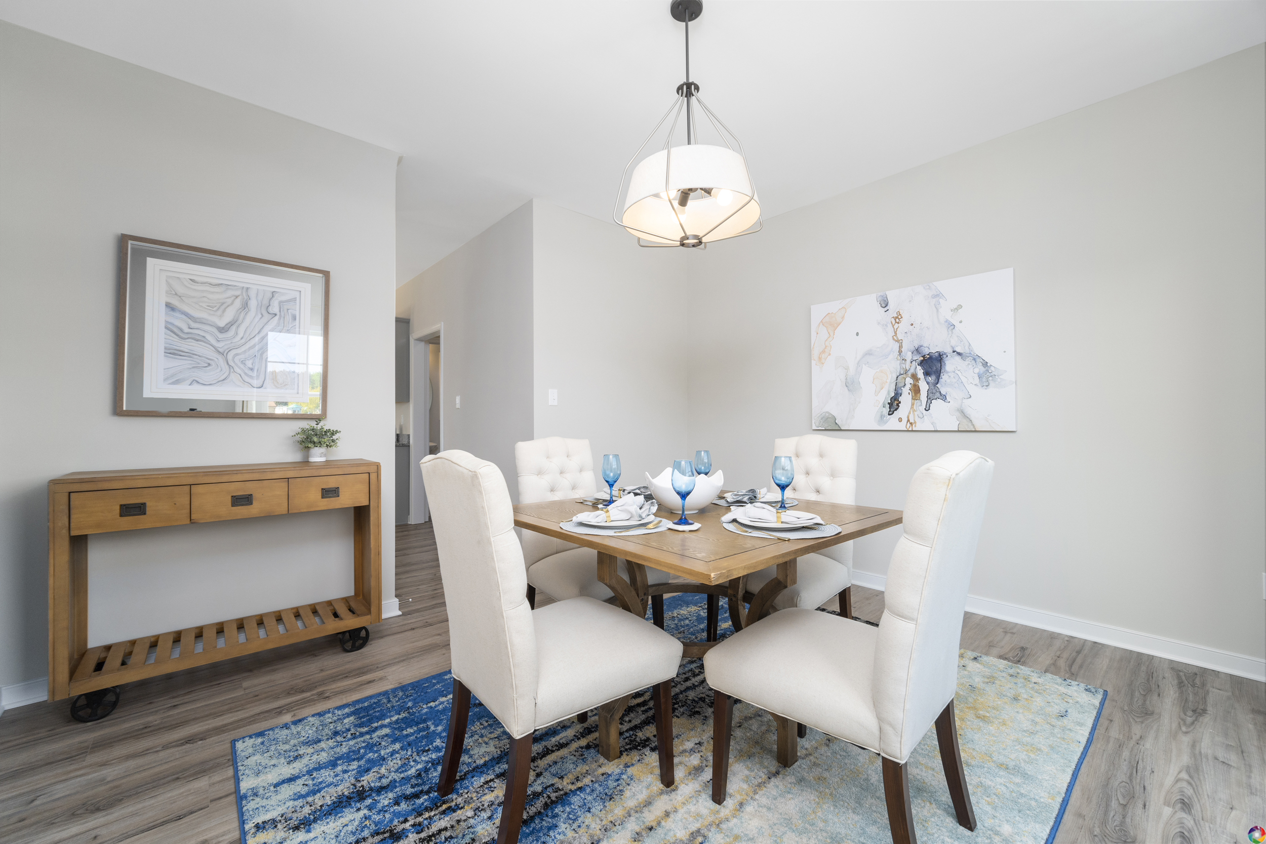 A dining room with a wooden table set for four, surrounded by upholstered chairs. The room features a sideboard, framed artwork on the walls, and a modern light fixture above the table.