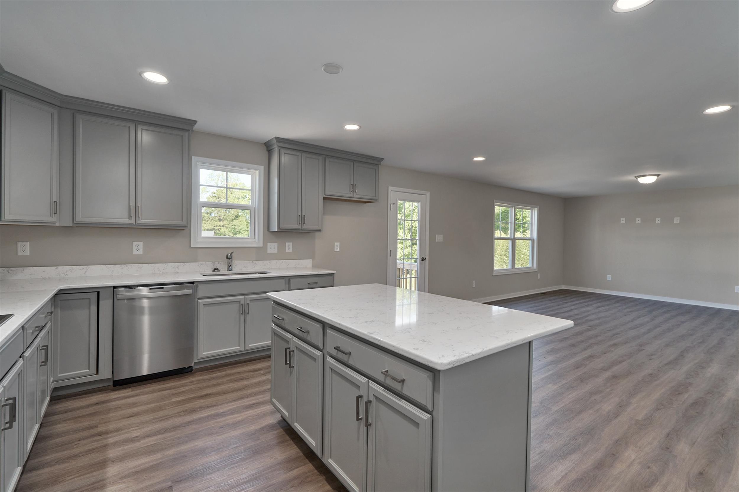 A modern kitchen with gray cabinets, stainless steel appliances, and a white marble island opens into a spacious living area with wood flooring and large windows.