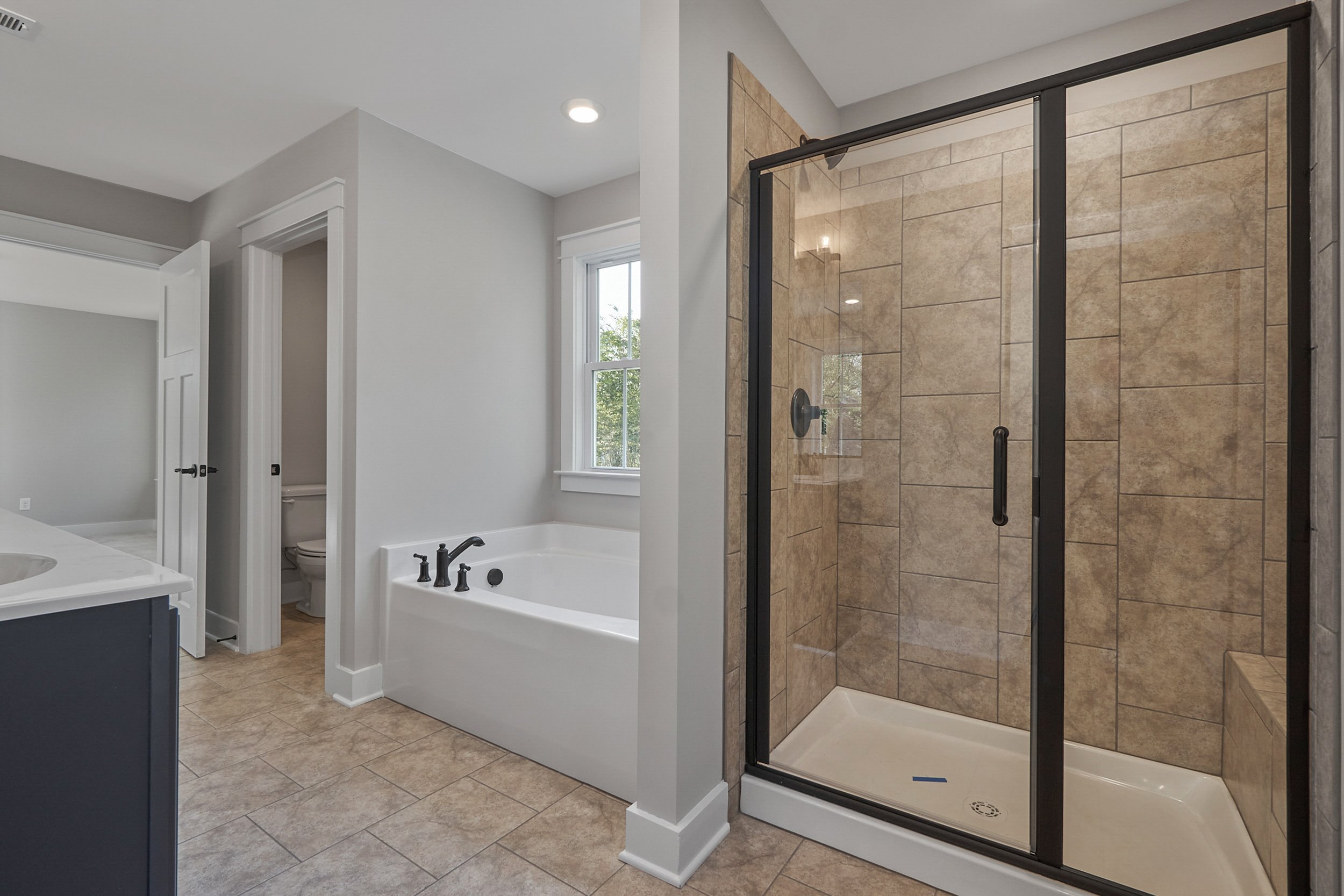 Modern bathroom featuring a glass-enclosed shower, a separate bathtub, a toilet in a small room, and a sink counter. The room has tiled floors and a window allowing natural light.