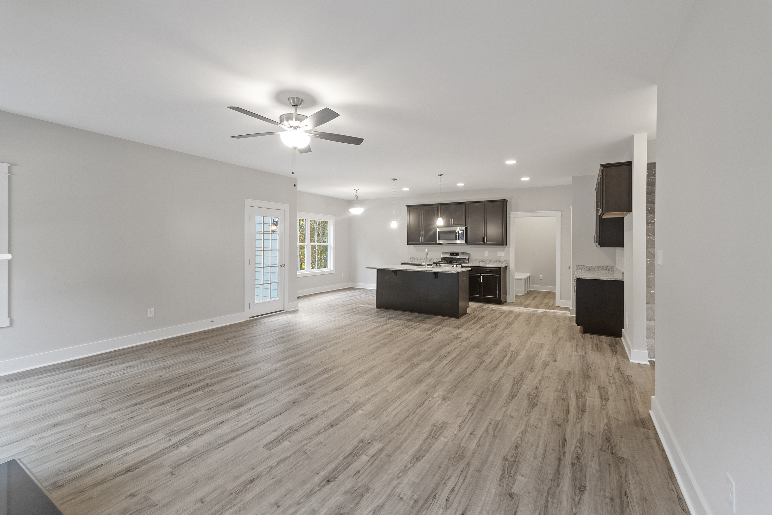 An open-concept kitchen and living room with wood floors, dark cabinetry, recessed lighting, ceiling fan, and French doors leading to an outdoor area.