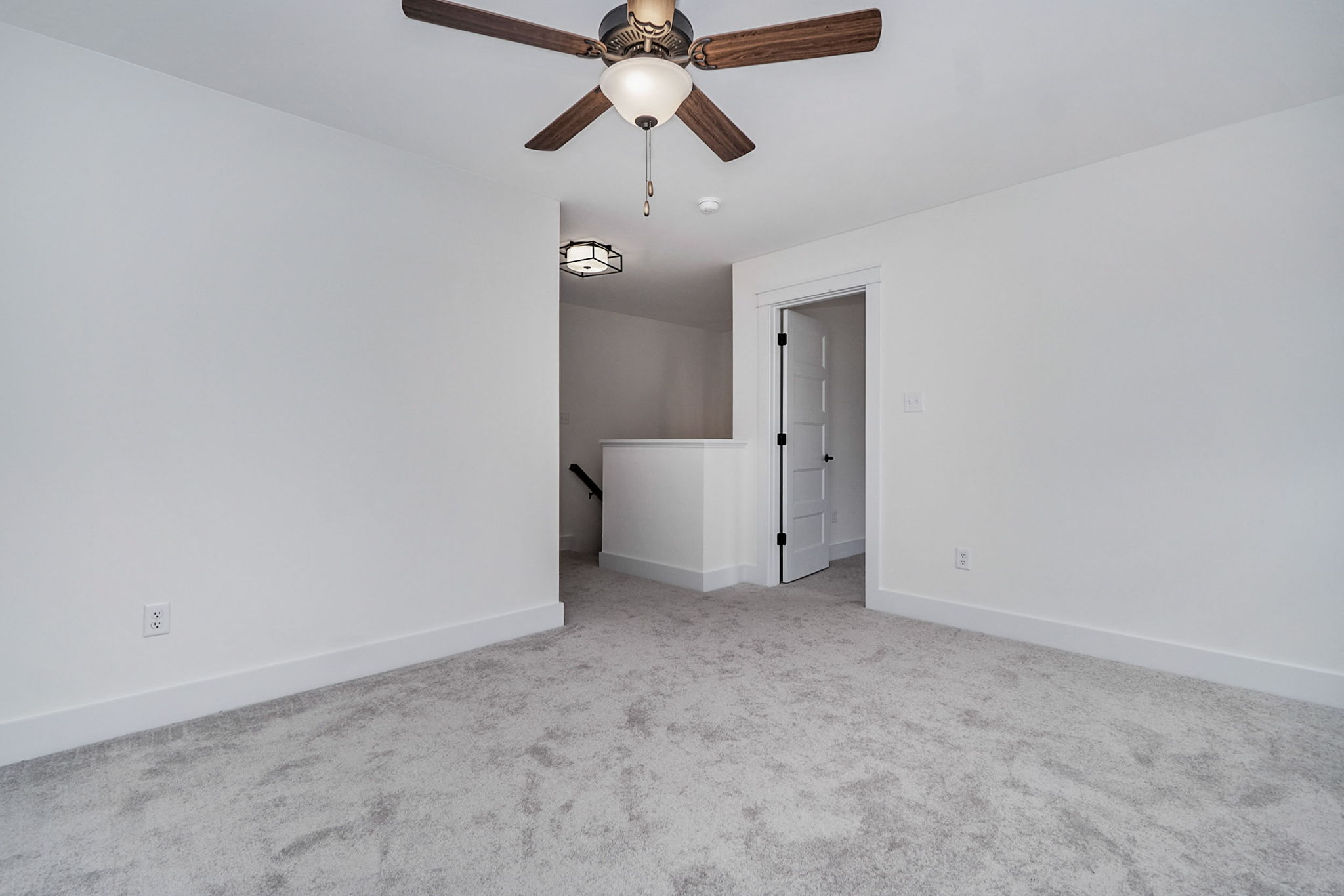 A room with light gray carpet, white walls, a ceiling fan with lights, and a white door partially open to a small hallway.