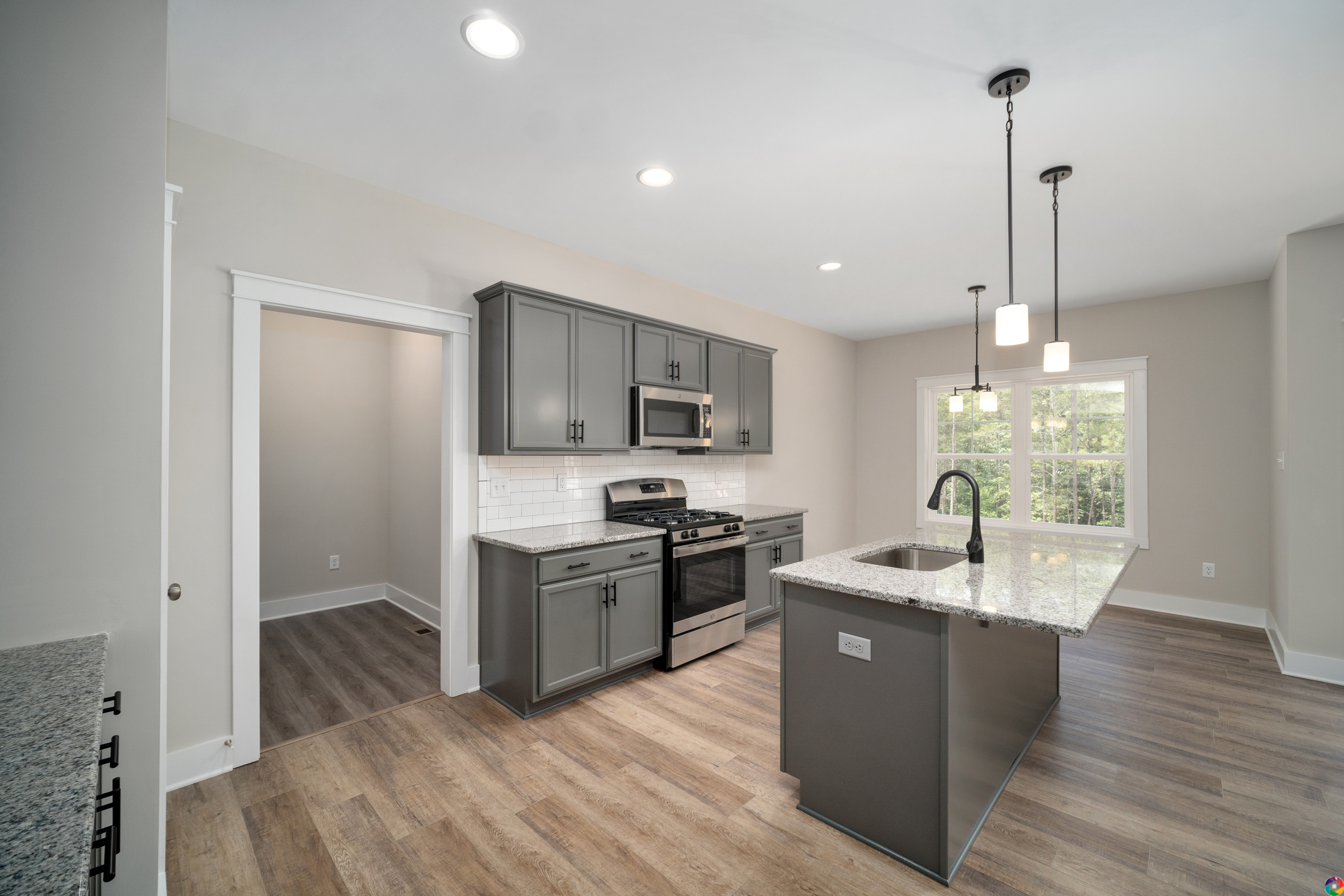 A modern kitchen with grey cabinets, white subway tile backsplash, stainless steel appliances, an island with a granite countertop, pendant lighting, and hardwood flooring.