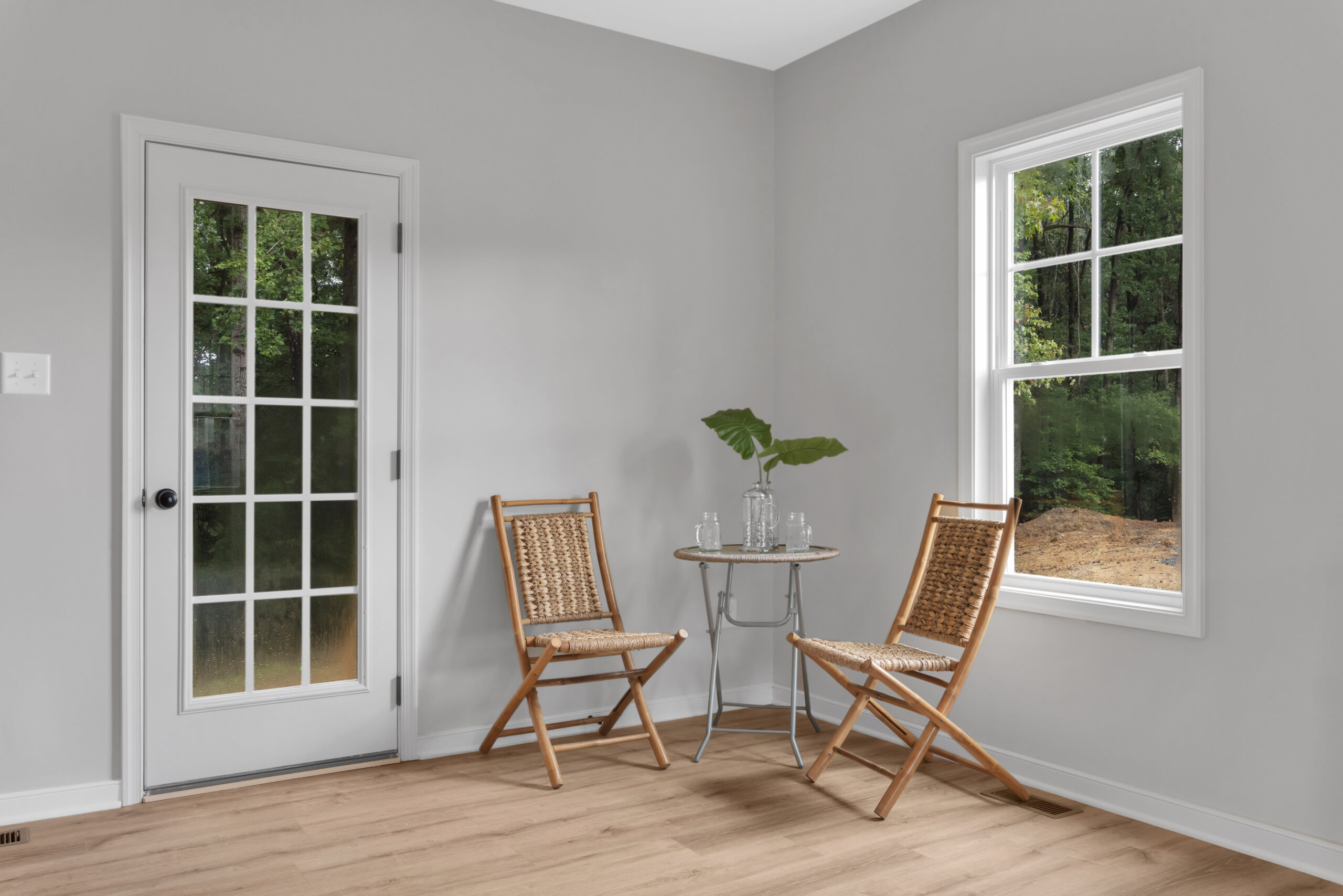A small room with light gray walls and a wooden floor features two wicker chairs and a small table with a glass pitcher and cups, near a window and a glass-panel door.