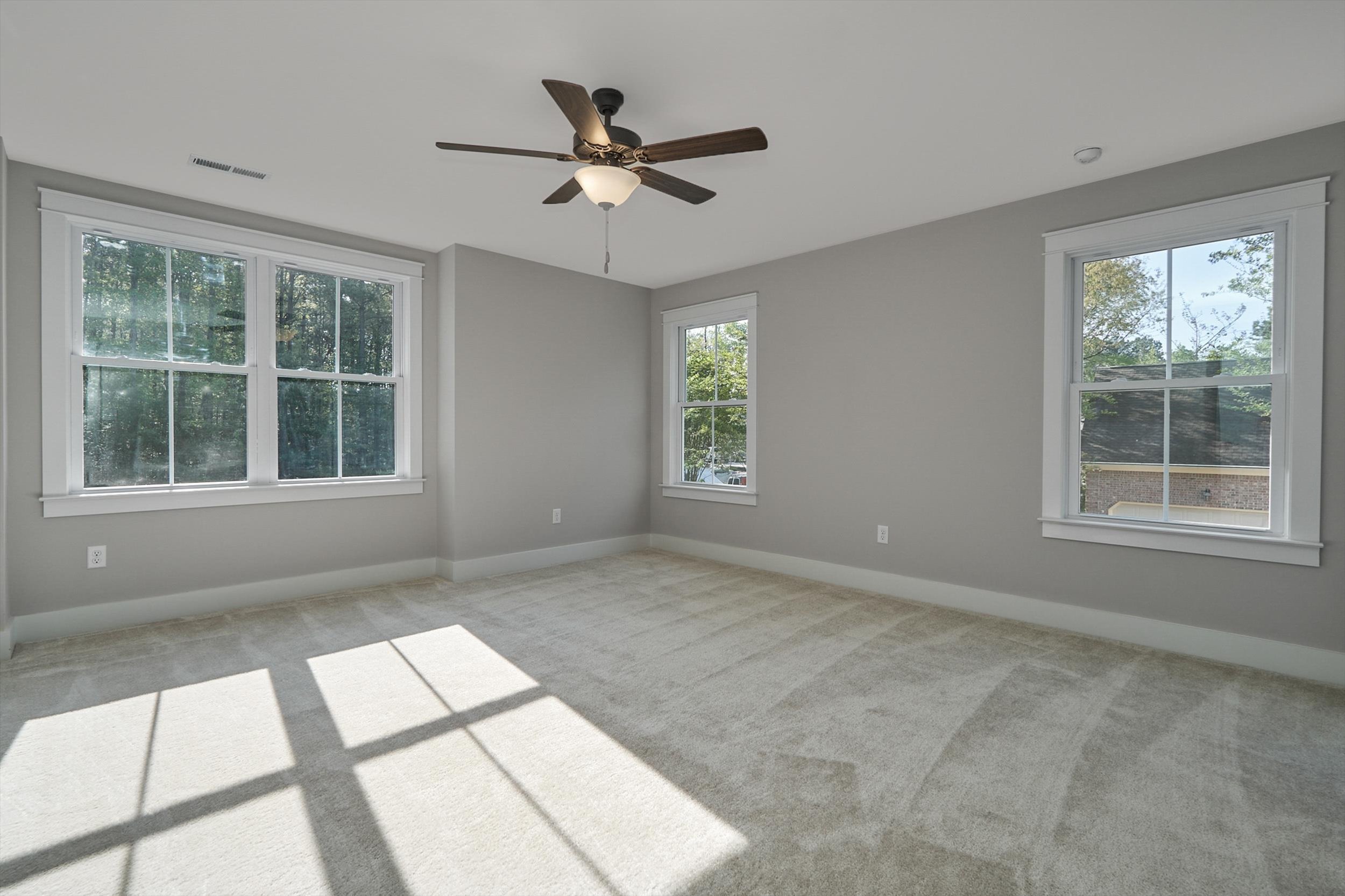 A bright, empty room with beige carpet, gray walls, a ceiling fan with a light, and three large windows letting in natural light.