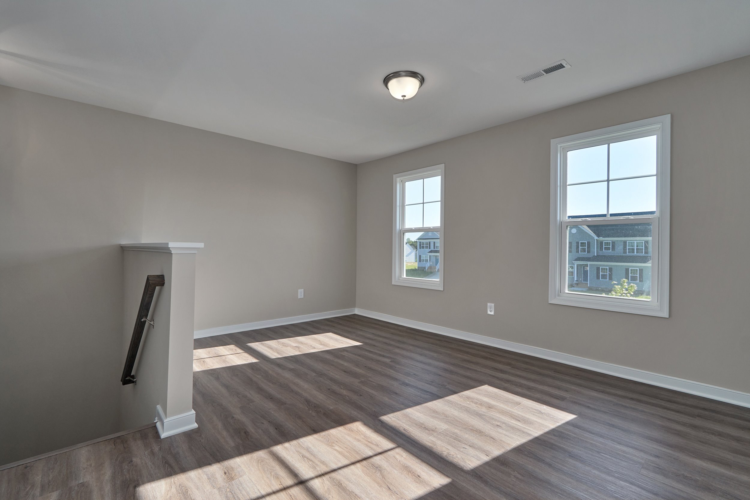 An empty room with light gray walls, two windows, and wood-look flooring; a small staircase is partially visible on the left.