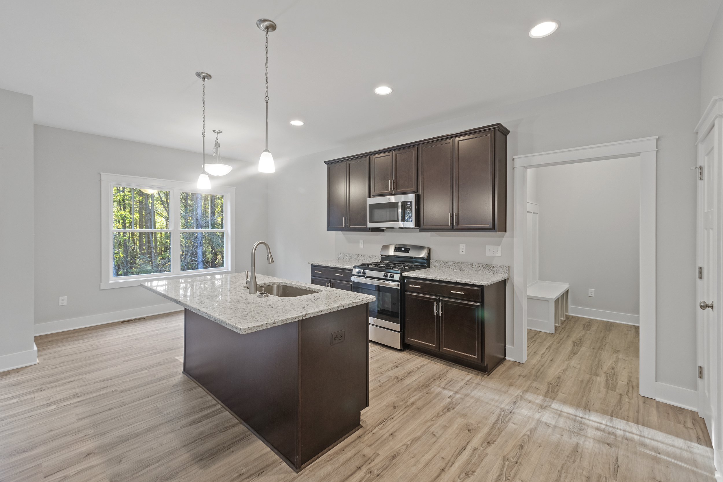 Modern kitchen with light wood flooring, an island with a granite countertop, pendant lights, dark cabinetry, stainless steel appliances, and a window providing natural light. Adjacent hallway visible.