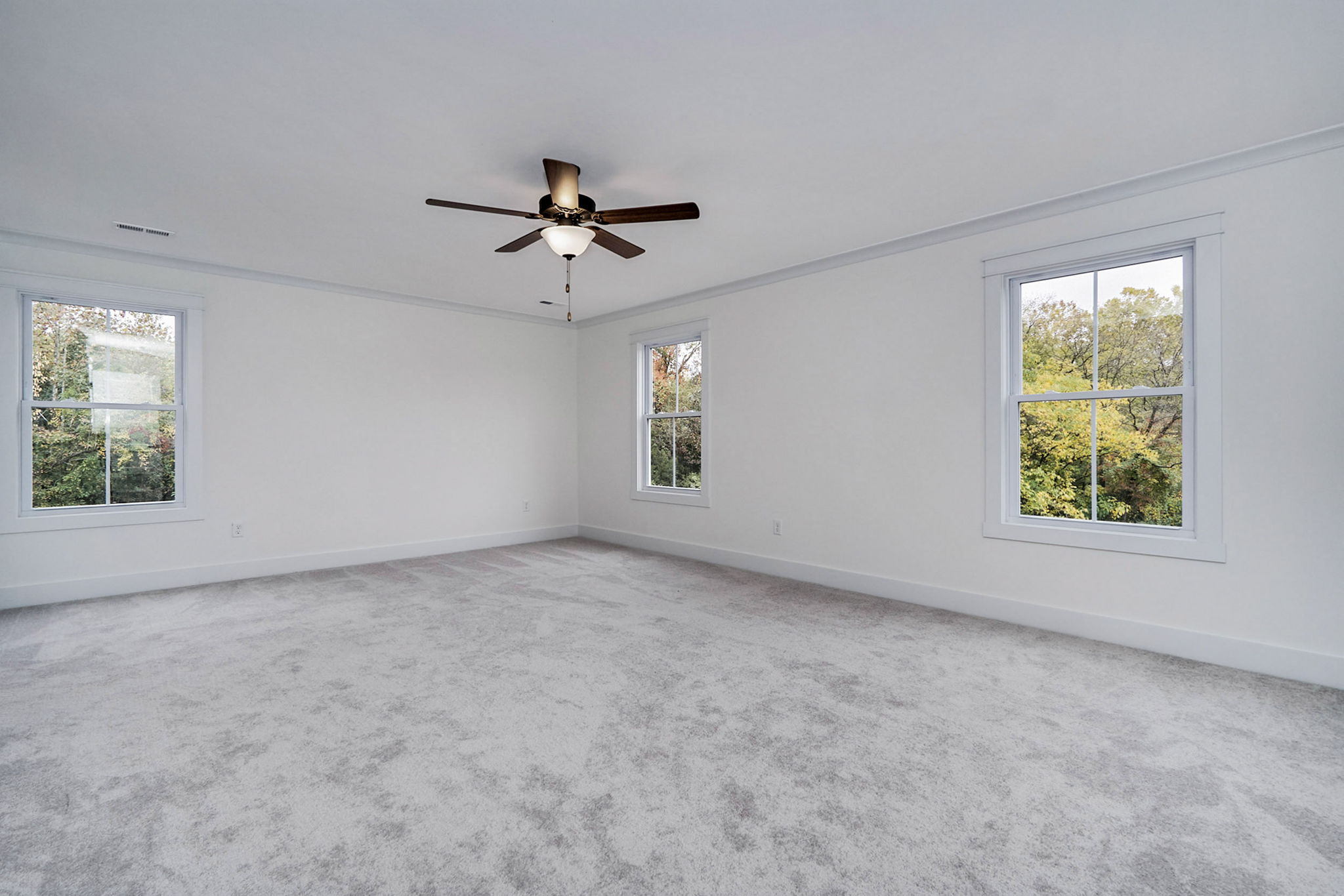 A spacious empty room with light-colored carpet, white walls, two windows, and a ceiling fan.
