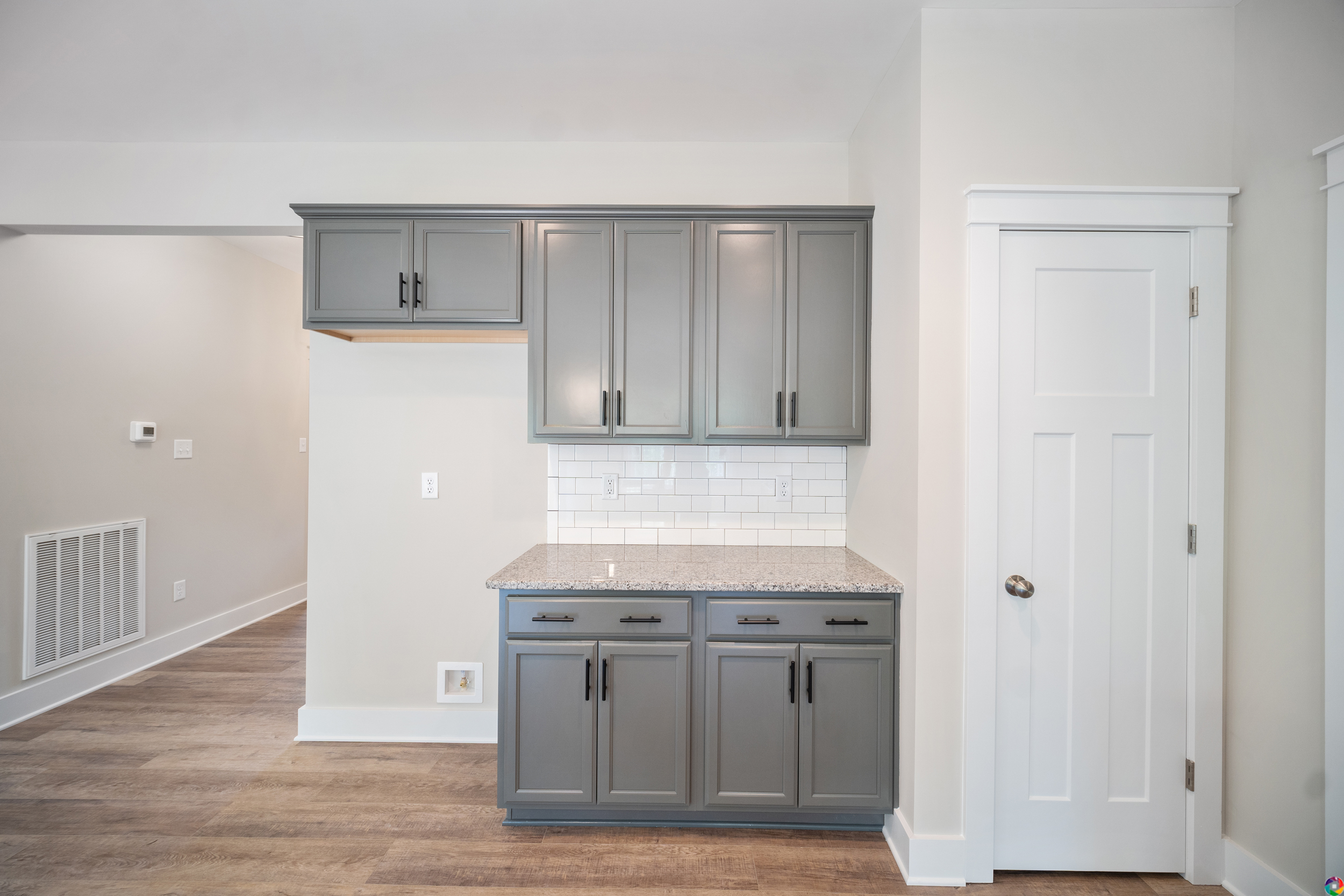 A kitchen area with gray cabinets, granite countertop, white subway tile backsplash, and wooden flooring. There is a white paneled door to the right.