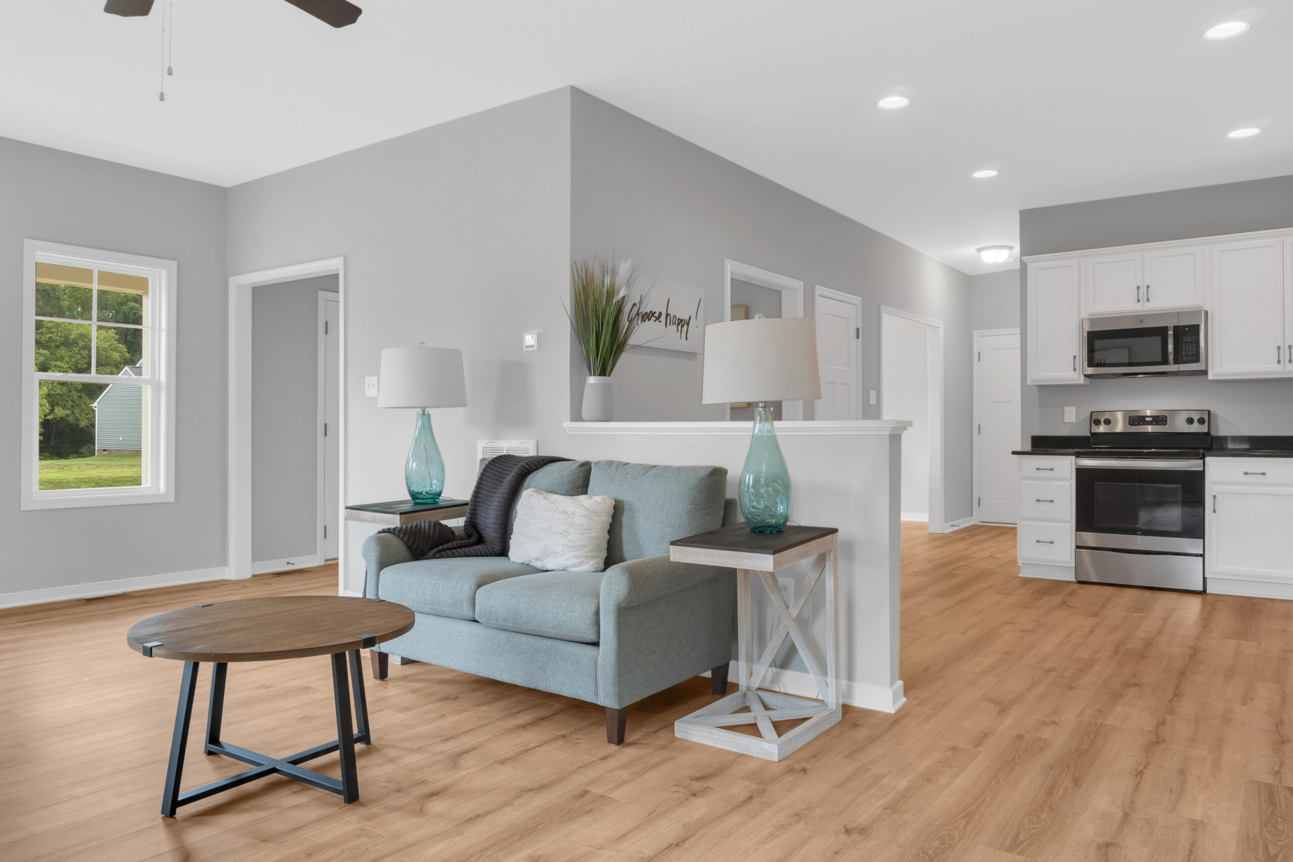 Modern living room connected to a kitchen with stainless steel appliances, featuring teal-blue sofa, wooden flooring, and gray walls.