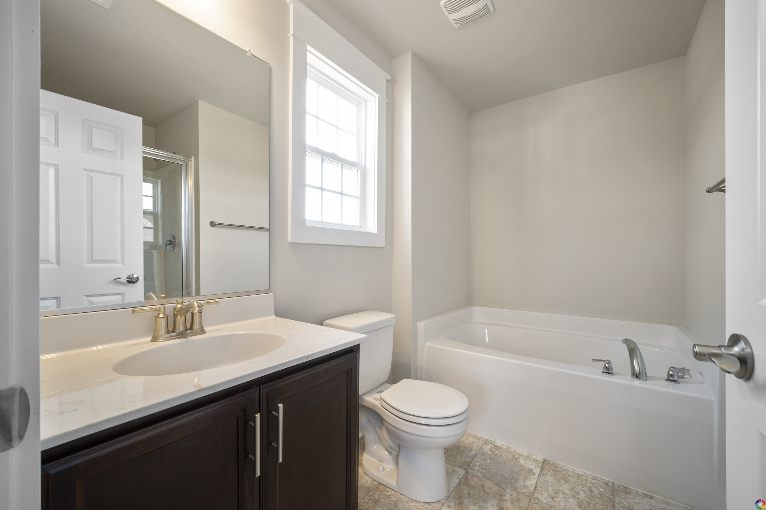 A clean, modern bathroom with a white bathtub, a toilet, a dark wood vanity with a sink and countertop, a large mirror, and a window above the tub.