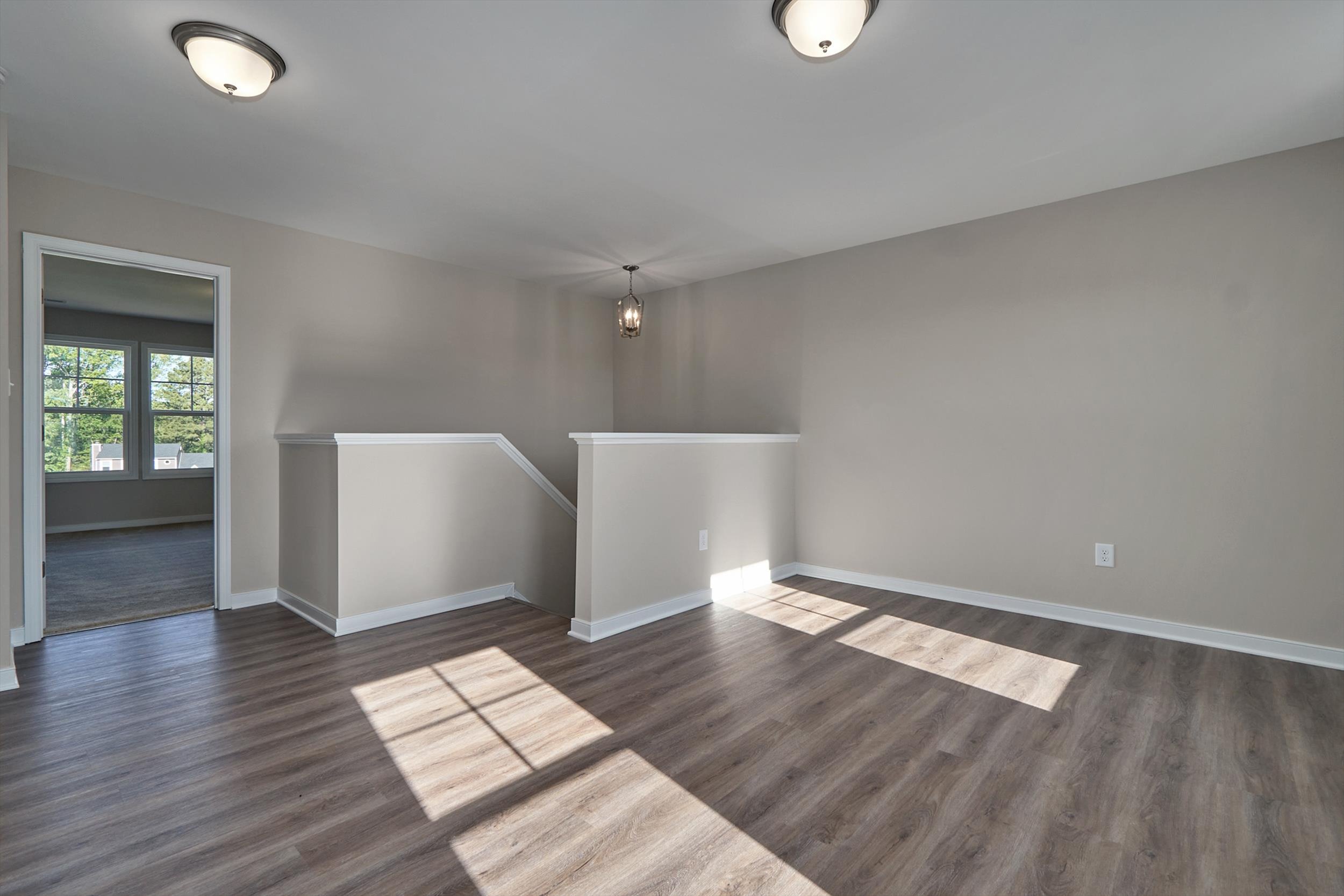 An empty room with wood flooring, beige walls, and two light fixtures on the ceiling. Adjacent is a stairway with a small landing area and a doorway leading to another room.