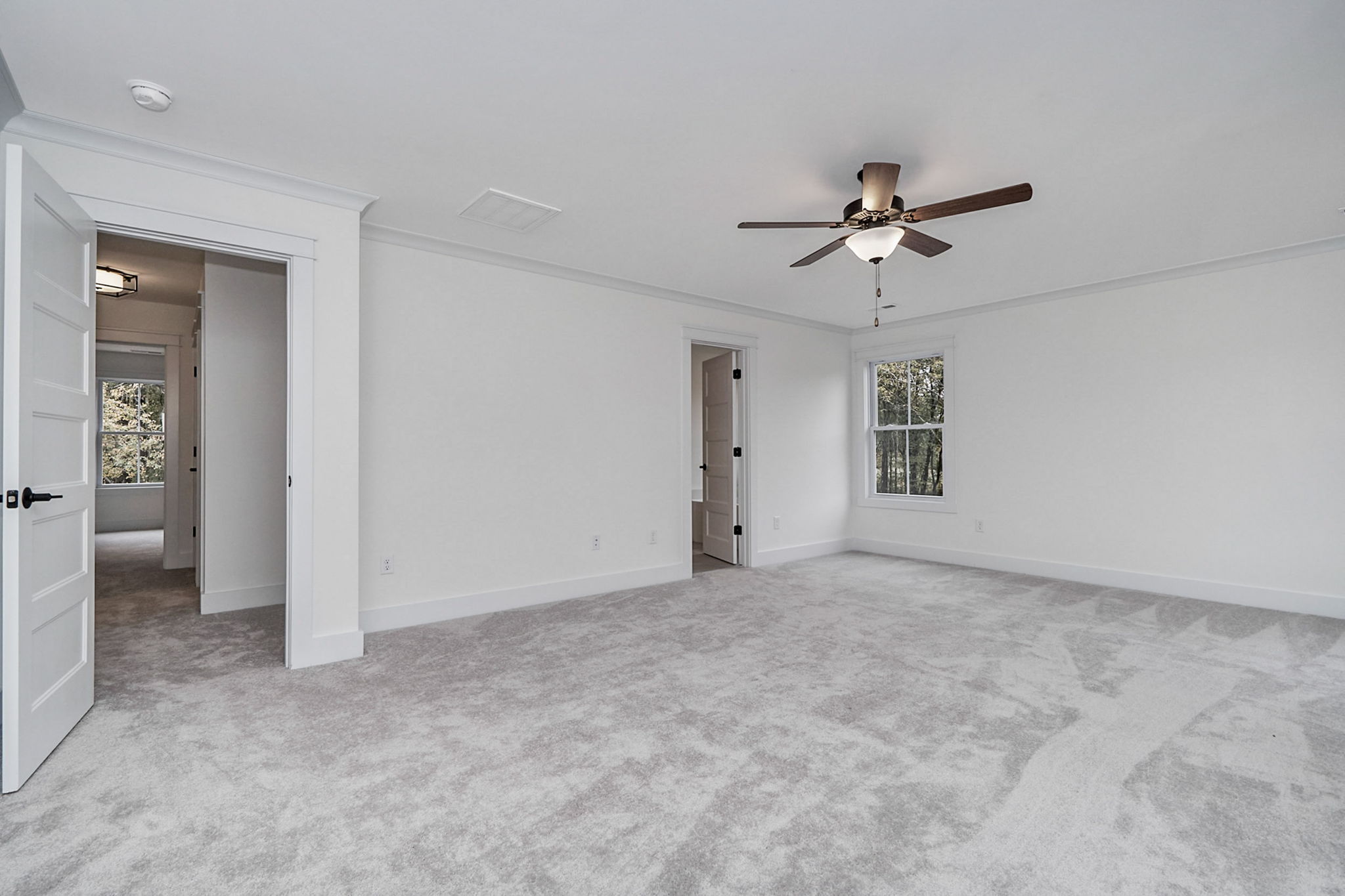Spacious empty room with light gray carpet, white walls, a ceiling fan, and open doorways leading to other rooms. A window provides natural light.