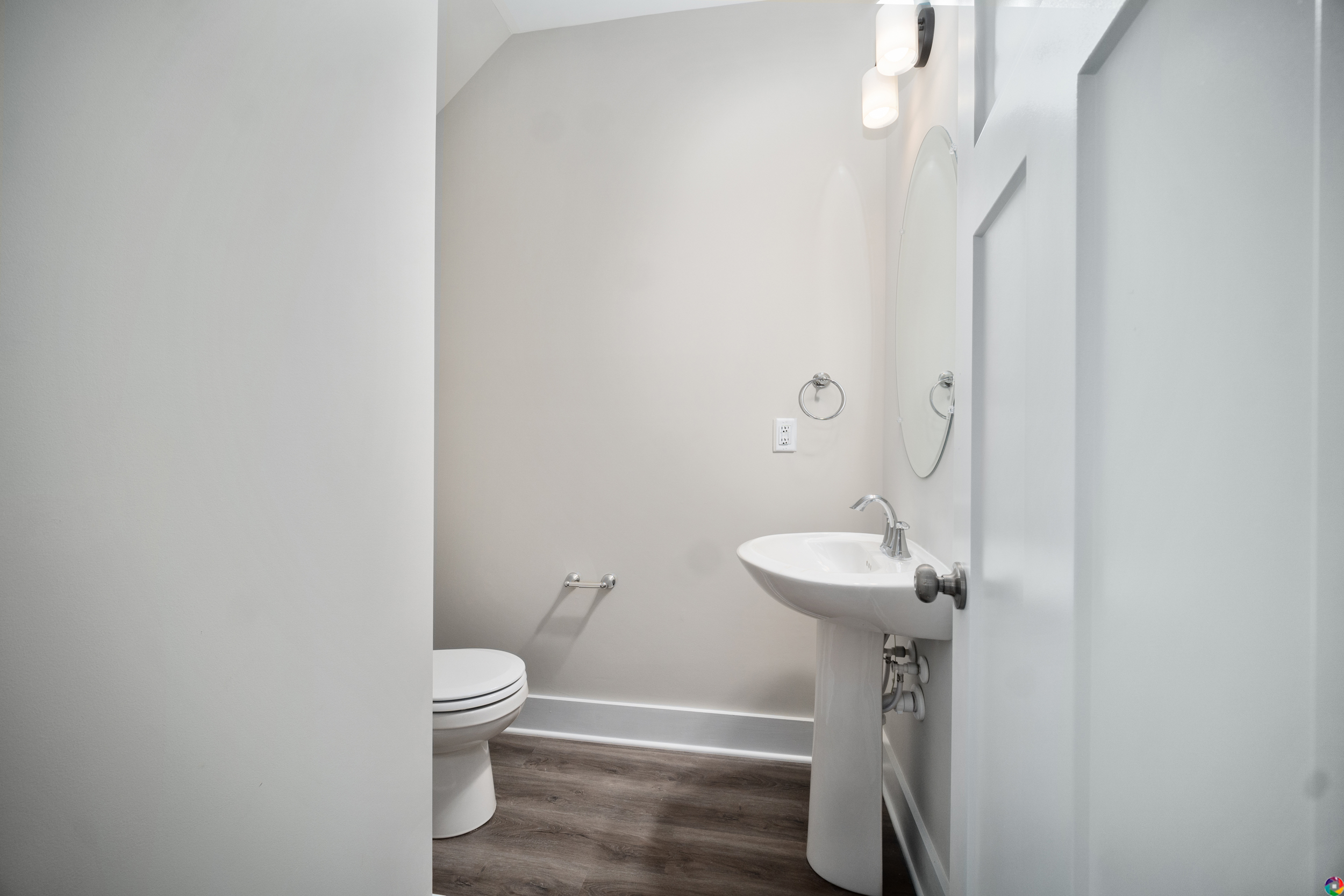 A small white bathroom with a toilet, pedestal sink, circular mirror, and wall-mounted light, featuring light gray walls and dark hardwood flooring.