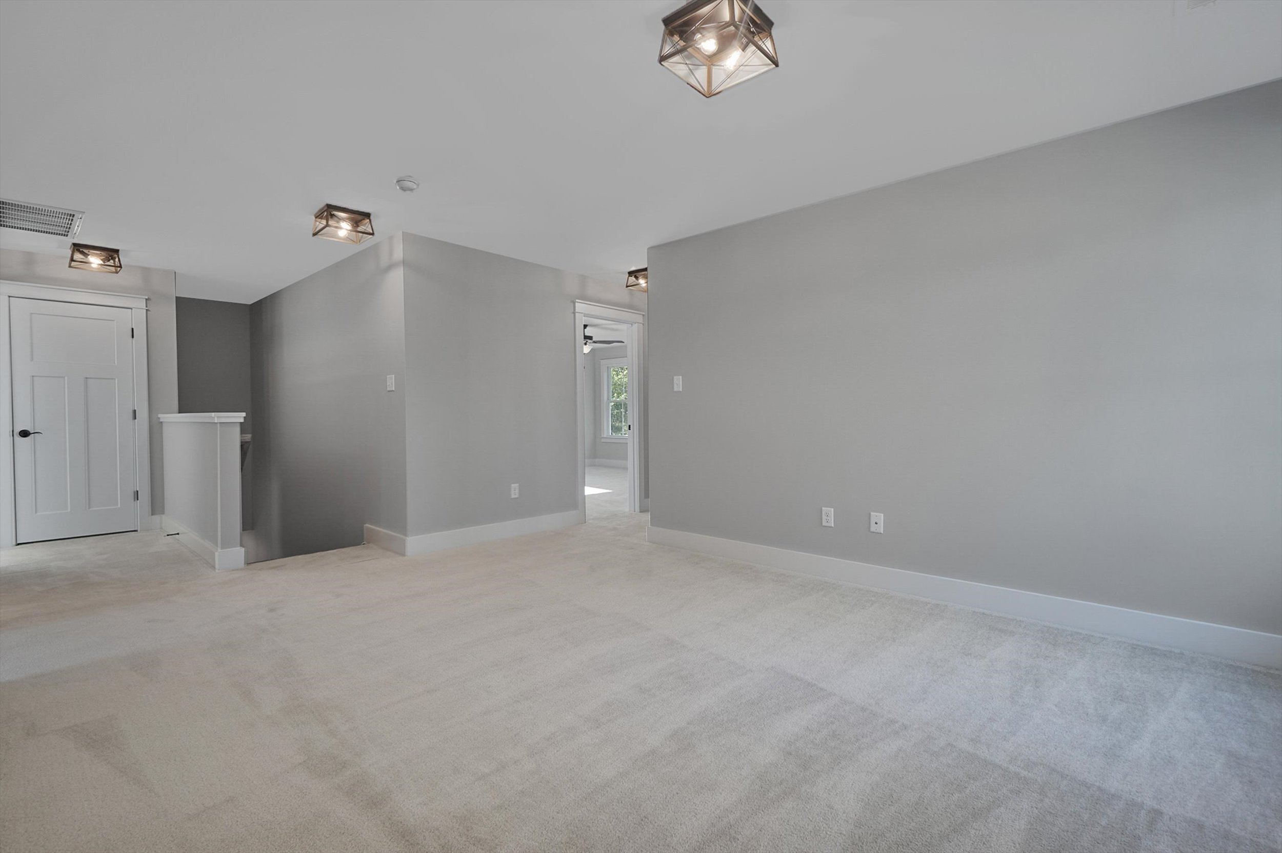 A spacious, carpeted room with grey walls, modern light fixtures, and a white door at the end of a short hallway. Natural light enters from a doorway on the right side.